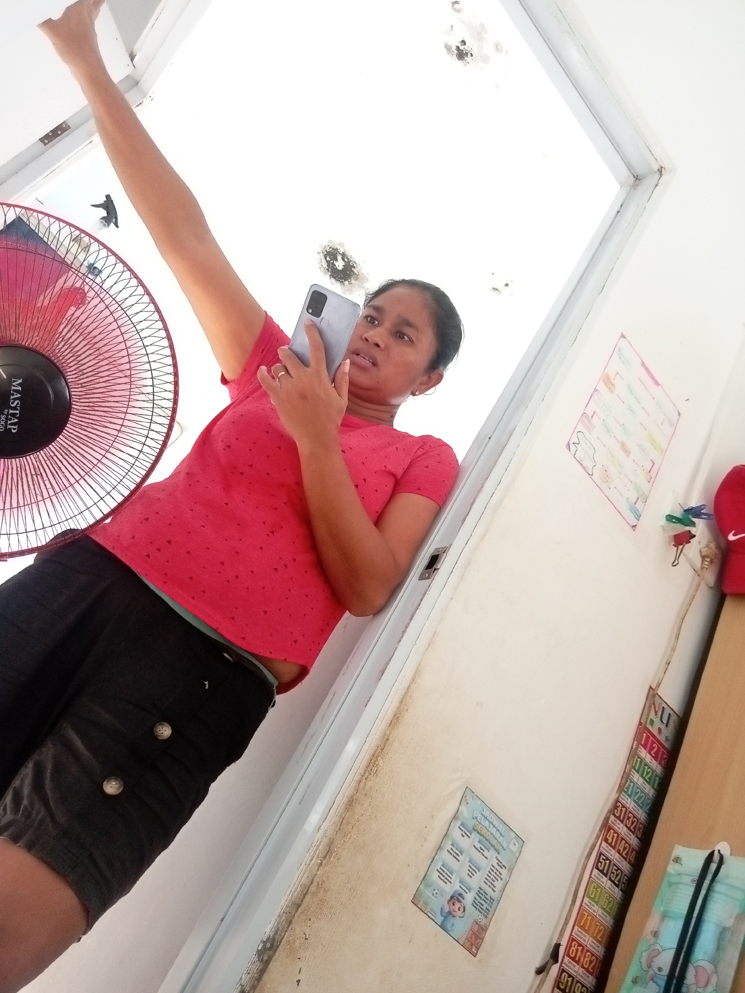 A low-angle medium shot shows a woman taking a selfie with a smartphone in a room. The woman, positioned in the lower right quadrant of the frame, is wearing a red t-shirt with a small geometric pattern and black shorts. She is leaning against a white wall and looking at the phone in her hand. Her left arm is raised and bent at the elbow, her hand holding the phone, which is pointed towards her face. A ring is visible on her left ring finger. To the left of the woman, a red electric fan with a black center is partially visible. The background consists of a white ceiling with some water stains and a white door frame. On the wall to the right of the woman, several colorful posters are taped, including one with a numbered grid and another with illustrations of children. A red object, possibly a hat, is also visible on the right side of the frame. The room appears to be a residential space. Based on the presence of a calendar with the letters "NU" which stands for Nahdlatul Ulama, a prominent Islamic organization in Indonesia, and typical home decor elements seen in Indonesian households, the location is likely a home in Batam, Indonesia.