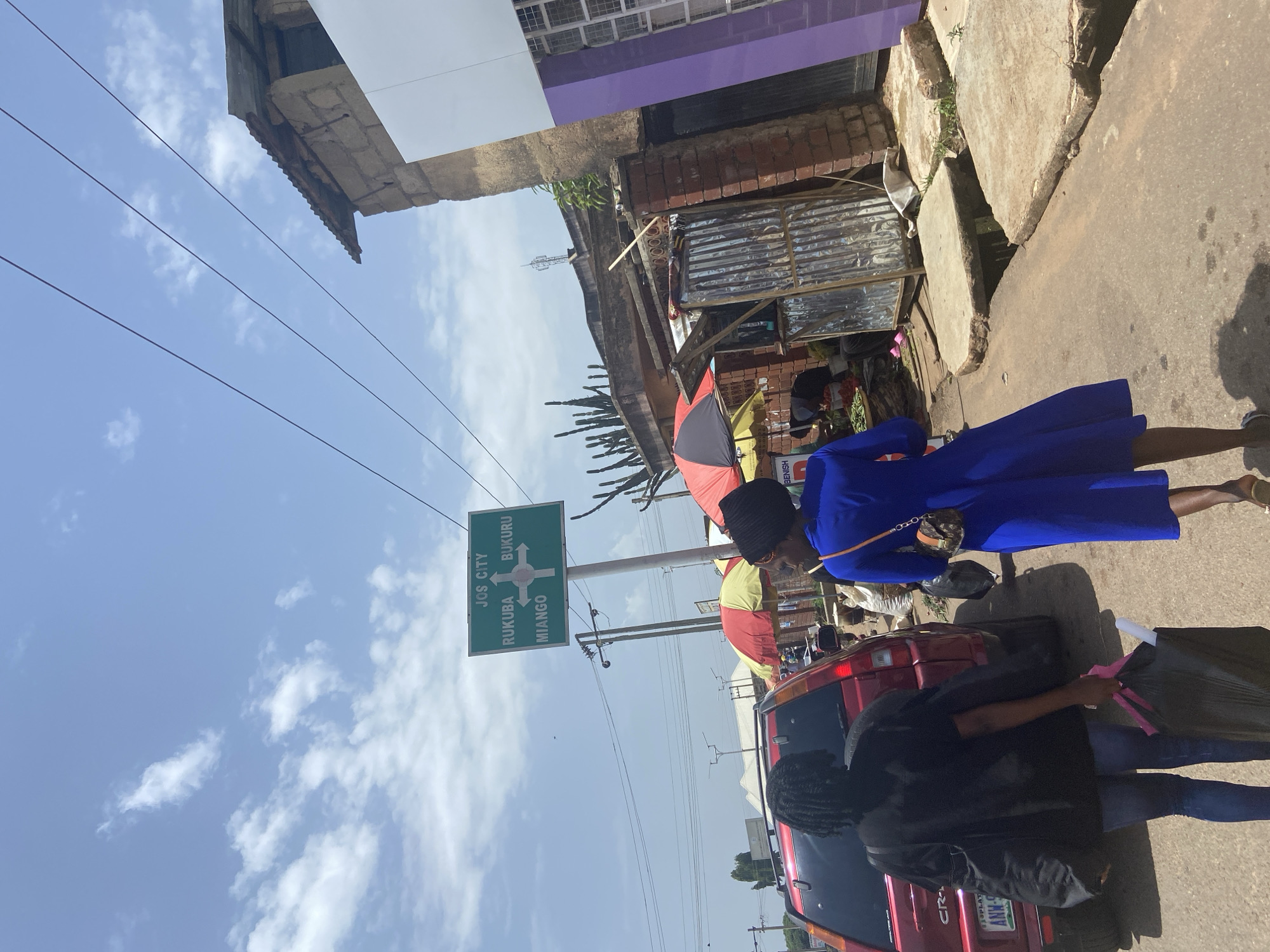 The image depicts a street scene in Jos, Nigeria, likely near a market or intersection given the presence of vendors and a directional signpost. In the foreground, two individuals are walking away from the camera. The person on the left is wearing a black top and blue jeans, carrying a large black bag in their left hand and a smaller black bag over their shoulder. The person on the right is wearing a vibrant blue dress and carrying a small purse.
A red SUV is parked to the left of the frame, with its license plate visible. Beyond the vehicles, a directional sign indicates "JOS CITY," with arrows pointing to "RUKUBA," "MIANGO," and "BUKURU." A market area with colorful umbrellas, stalls constructed from corrugated metal, and visible produce is situated to the right. The environment appears to be an urban setting with buildings made of concrete blocks and corrugated iron roofing, along with overhead power lines. The sky is bright blue with scattered clouds, suggesting daytime. The ground is paved, with a section of open drainage visible in the middle right of the image. The overall impression is a bustling, everyday scene in a Nigerian city.