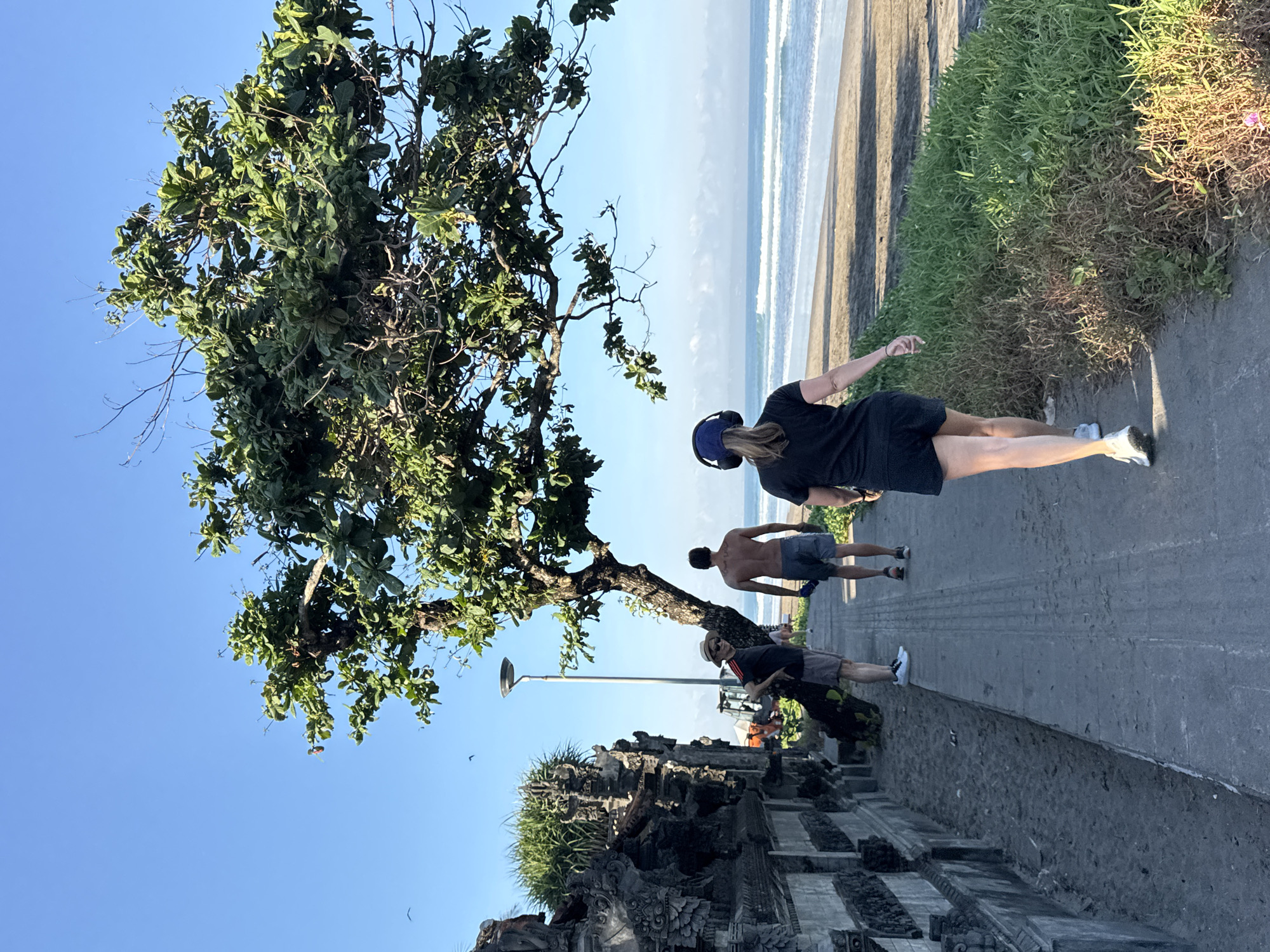 The image shows a partially obstructed view of Kuta Beach in Kuta, Indonesia. Four people are visible on a paved walkway; a woman in a black dress is walking, two men are walking behind her, and a fourth man is seated. The beach is to the right of the walkway, separated by vegetation. Waves can be seen along the shoreline. To the left of the walkway is a stone structure, possibly part of a temple. A large tree is above the walkway. A street light is visible behind the tree. The sky is blue and clear.