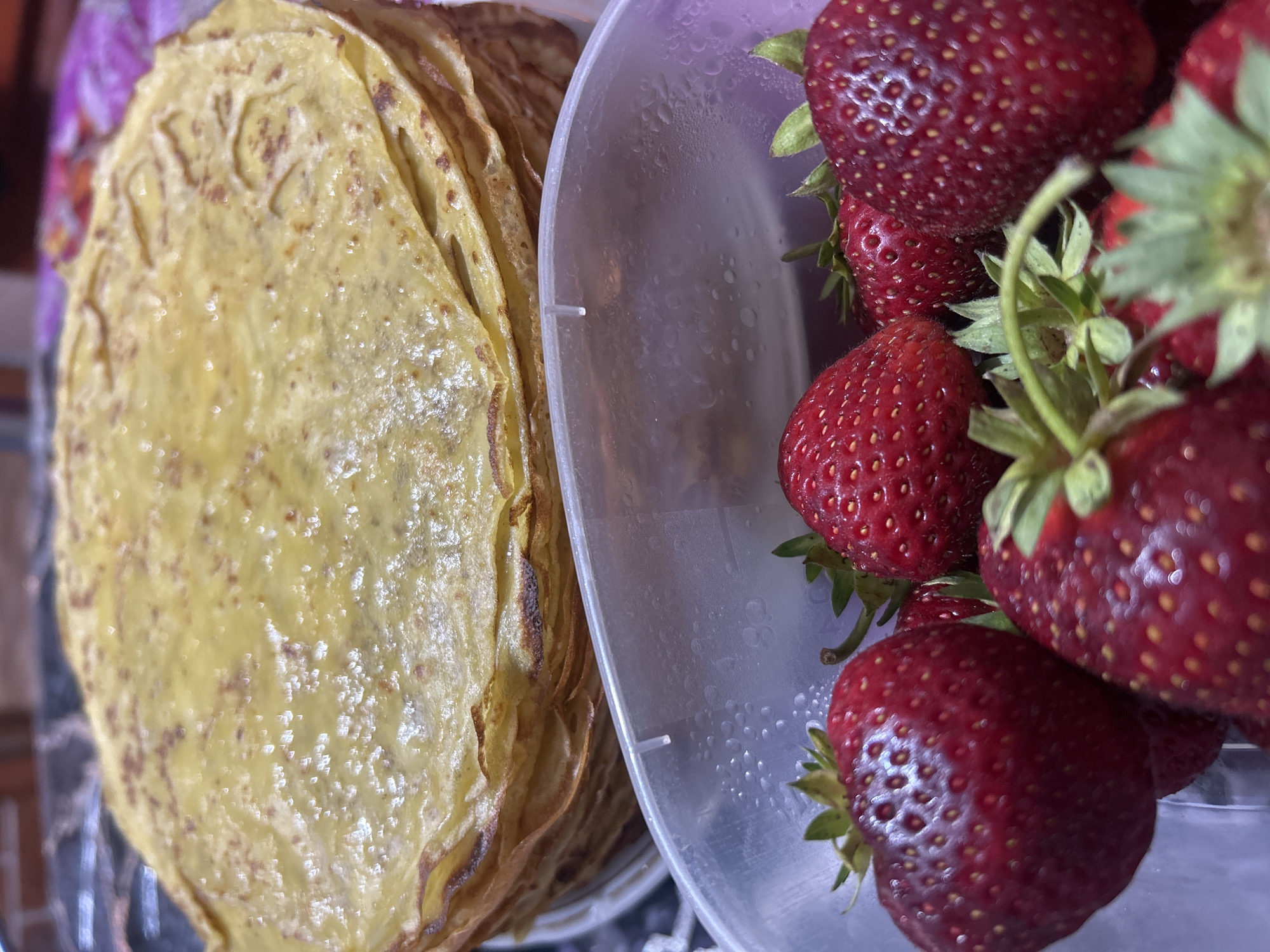 A stack of golden crepes sits behind a clear plastic container filled with fresh strawberries. The crepes are thin and layered, with some browning visible on the edges. The strawberries are ripe and red, with green stems and leaves. The container is partially filled with what appears to be condensation. The background is blurred, but hints of a dark, possibly marble countertop and a brick wall can be discerned. This scene is not specific to Gogino, Russia, or any other city.