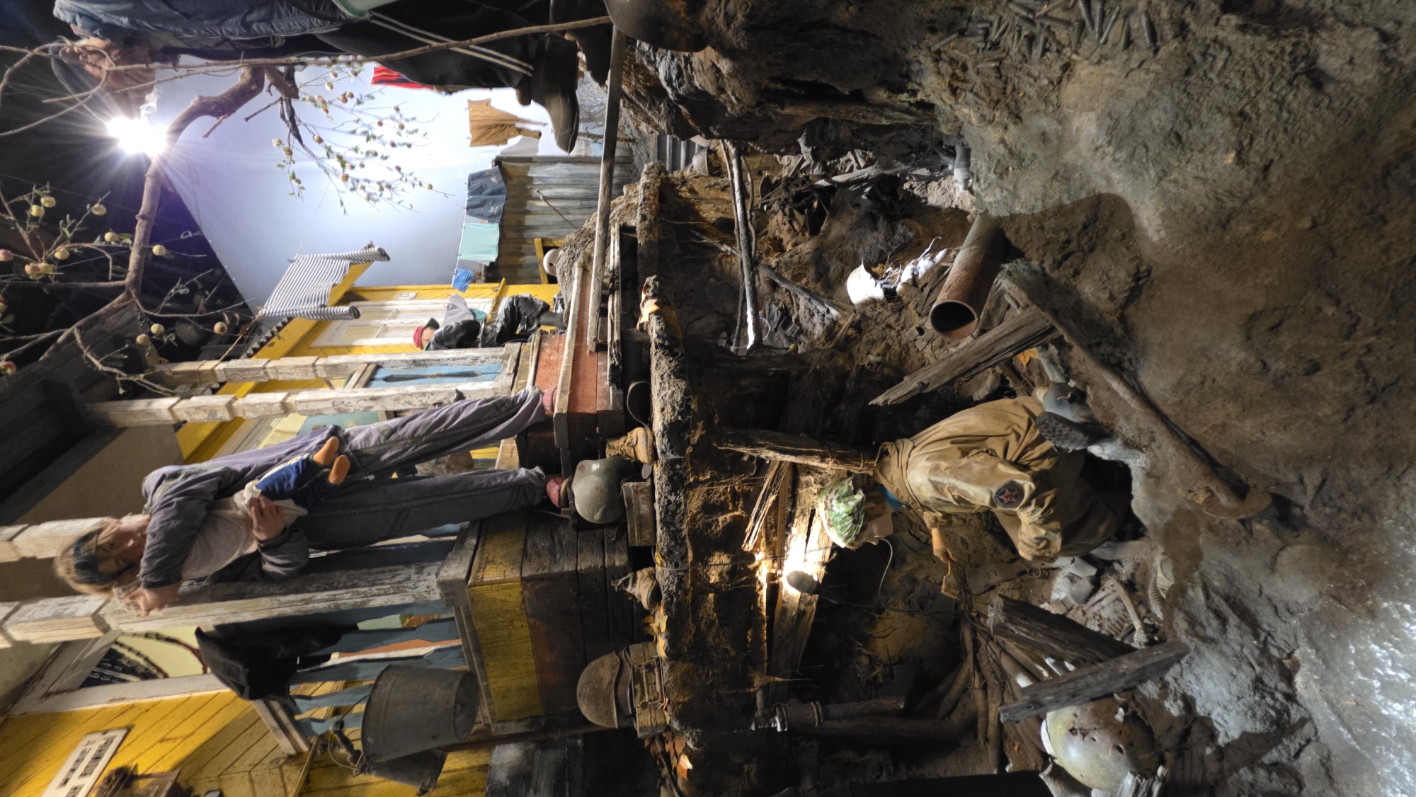 A dimly lit, multi-level display depicts a scene that transitions from a bombed-out city to an underground bunker.
On the left, a yellow building structure with windows and draped items is visible. A woman is lying horizontally, holding a doll in her arms, possibly representing a mother and child in a shelter. Above, there is a simulated sky with artificial lighting and a tree branch with hanging fruits.
The right side of the image shows a man, a soldier in a military uniform in what appears to be a dark, rough-hewn bunker or underground shelter. The walls are rocky and earthen, with miscellaneous objects scattered throughout, possibly indicating a makeshift living space during conflict. The scene is filled with artifacts, debris, and props to create a sense of historical realism.