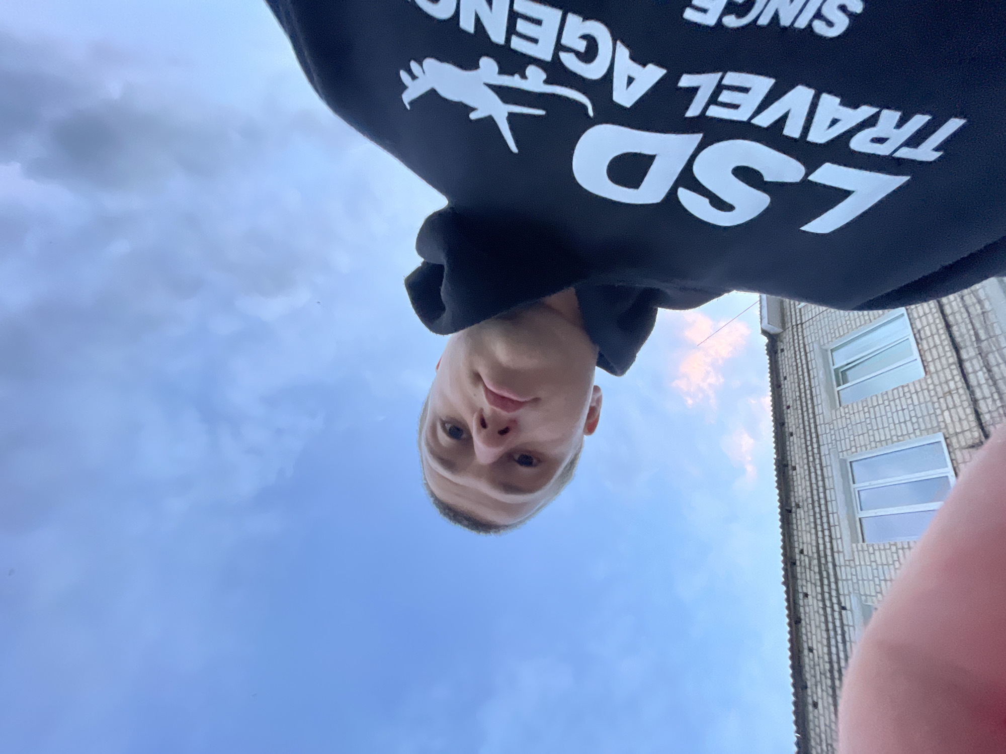 This image presents a unique, low-angle perspective, looking upwards, with the main subject appearing upside down against a cloudy sky and a building. The central figure is a young man, whose face is visible at the bottom center of the frame. He has light skin, short light hair, and looks directly at the camera with a neutral expression. He is wearing a black hooded sweatshirt.
The sweatshirt features white text and a graphic. From the viewer's perspective, the text appears upside down, reading "LSD TRAVEL AGENCY" and "SINCE". Above the words "TRAVEL AGENCY" is a white silhouette of a person, possibly a skier or snowboarder, captured in mid-air.
To the right side of the image, a light-colored brick building with multiple white-framed windows is partially visible. A portion of what appears to be the photographer's arm or hand can be seen in the bottom right corner, slightly obscuring the building. The majority of the background is a sky filled with greyish-blue clouds, indicating an overcast or partly cloudy day. There are faint hints of pinkish-orange hues in the clouds near the building, suggesting either early morning or late afternoon light.
The image appears to be a selfie taken from an unconventional angle, likely with the camera held below the subject's face and pointed upwards, or rotated after capture. The setting is outdoors in Красные Баки, Russia, presumably near a residential building.