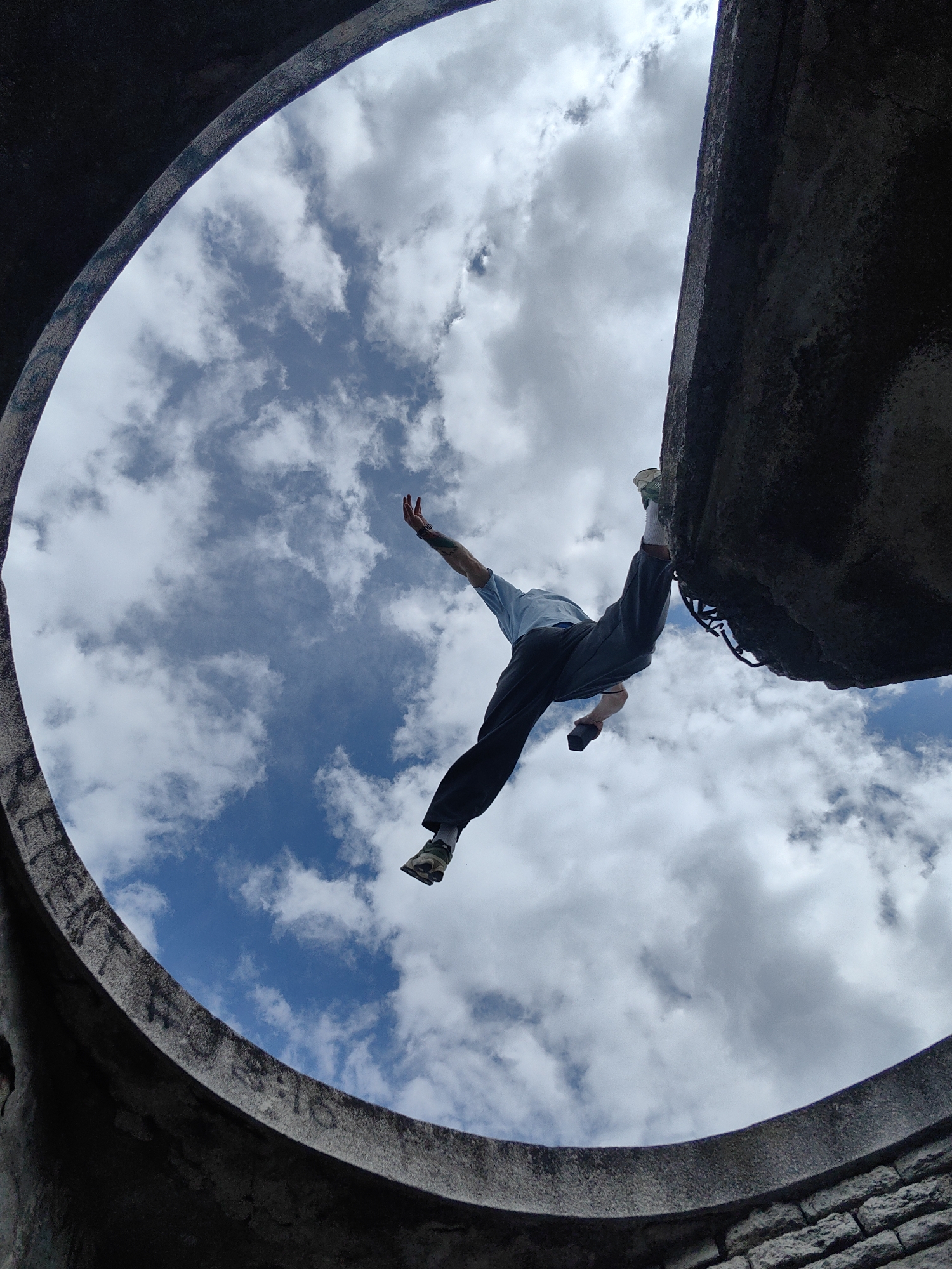 This low-angle photograph, taken from inside a circular concrete structure, looks up towards a bright, cloudy sky. The main subject is a person, likely male, captured in a dynamic, suspended pose against the backdrop of the sky. He is wearing a light blue t-shirt, dark trousers, and athletic shoes. One arm, which displays tattoos, is outstretched towards the upper left, while his body is angled, and his right leg is bent, with his foot seemingly balanced on a dark, rough ledge forming the edge of the circular opening on the right side of the frame. This pose suggests he is engaged in climbing, parkour, or a similar acrobatic activity, either entering or exiting the structure.
The setting is an opening in a massive, dark grey concrete or stone structure, possibly a well, tunnel, or ruin, located in Varna, Bulgaria. The inner curve of the structure is visible, framing the sky, with its surface appearing rough and worn. Faint graffiti, including the word "FREE" and other illegible letters and numbers, can be seen along the left side of the circular edge. The sky is a vivid blue with scattered white, fluffy clouds, indicating a clear, bright daytime. The perspective emphasizes the scale of the structure and the daring nature of the person's action, creating a sense of movement and adventure.