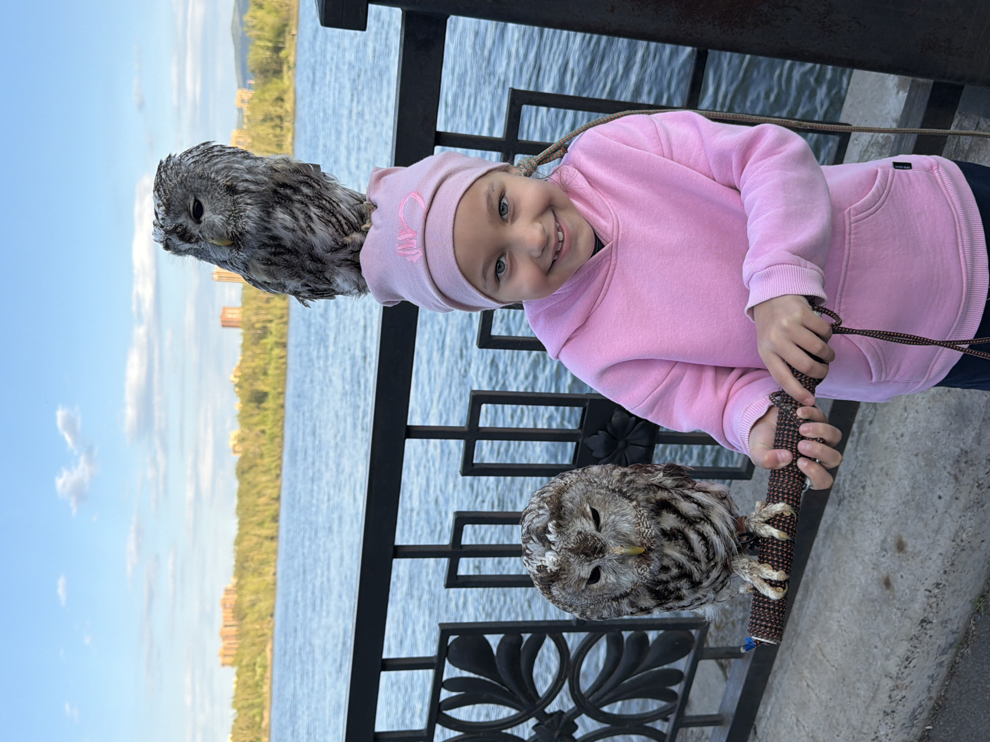 A young girl in a pink hooded sweatshirt and a matching pink hat stands on what appears to be a waterfront promenade. She is smiling and looking directly at the camera. She is holding a perch with two owls. One owl is perched on the top of the perch, near her head, and the other owl is perched near her hands at the bottom of the perch. A black ornate railing separates the girl from the water, which reflects a strip of green trees and distant buildings under a pale blue sky with light clouds. This scene is located in Krasnoyarsk, Russia.