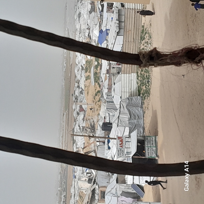 The image captures an expansive, densely populated informal settlement in Khan Yunis, Palestinian Territory, under an overcast or hazy sky. The ground throughout the visible area consists of sandy dirt with intermittent patches of sparse green vegetation.
The settlement is characterized by numerous makeshift shelters, predominantly constructed from white and light grey tarpaulins draped over various frames; some are dome-shaped, resembling yurts, while others are rectangular. In the mid-ground, a small, open-fronted kiosk with a white canopy stands on a dirt path. The kiosk's signage displays "المجد" (Al-Majd) in Arabic, along with other Arabic text. A black water tank is visible atop one shelter, and a utility pole with wires is positioned near the kiosk. A light-colored corrugated metal fence runs across the mid-right. A dark, rectangular object, possibly a solar panel, leans against a tent on the left. In the distant background, the settlement extends into a flatter, lighter-colored landscape under a uniform atmospheric haze.
Two large, dark, out-of-focus vertical objects, possibly tree trunks or poles, partially frame the foreground on the left and right. The right-side object exhibits tangled fibrous material at its base.
Three distinct groups of people are present:
1. A person in dark clothing walks from left to right on a dirt path in the left mid-ground.
2. A person wearing a long, dark robe stands with their back mostly to the viewer, facing right, near the corrugated fence in the right mid-ground.
3. Two small figures, one in a blue top and the other in a dark top, are partially visible in the bottom-right corner, moving away from the camera.
A figure is also partially visible behind the counter of the kiosk. The bottom-left corner of the image contains a "Galaxy A14" watermark.