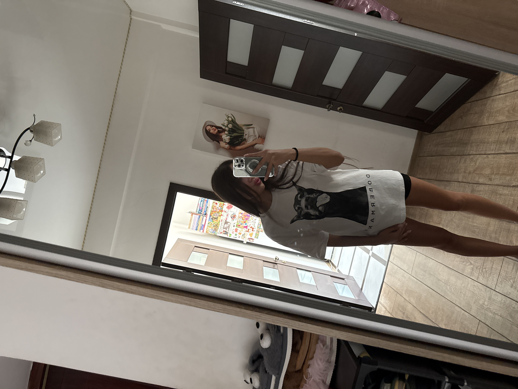 A young woman is taking a mirror selfie in what appears to be a hallway or entryway of a residential property, possibly located in Chișinău, Moldova, given the context. She is wearing a white t-shirt with a black graphic of a dog's face and the word "DOBERMAN" below it, and black shorts. Her hair is long and brown, and she is holding a smartphone in her right hand to take the picture. In the reflection, we can see a light-colored wall with a painting of a woman holding flowers, and a dark brown door with several rectangular frosted glass panels. To the left of the woman, also reflected, is a tall mirror frame, a shoe rack with grey fuzzy slippers, and another door. The flooring is made of light-colored wooden planks. The lighting suggests an indoor setting during the daytime.