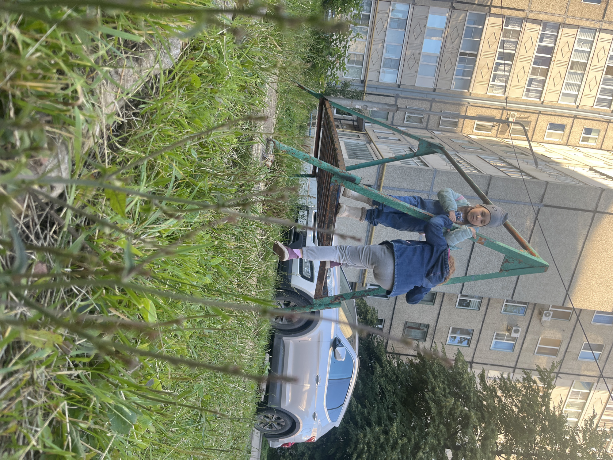 The image depicts two young children interacting on a rusty green metal playground structure in front of a grey, multi-story apartment building. The perspective is from a low angle, looking upwards, with lush green grass and plants in the foreground, partially obscuring the scene. The building has many windows and appears to be residential. A white SUV is parked on the right side of the frame, with its rear visible.
One child, wearing a grey cap, blue jacket, and grey pants, is lying on the metal frame. Another child, wearing a light blue and grey jacket, grey pants, and white socks, is also on the structure, appearing to be interacting with the first child. The children's clothing suggests a cool weather day.
The scene appears to be set in an urban environment, likely a residential courtyard, in Novocheboksarsk, Russia. The time of day seems to be afternoon, given the lighting. The overall mood is casual and playful, with the children engaged in an activity on the playground. No visible text is discernible in the image, except for what might be a license plate on the back of the car, but it is too blurry to read.