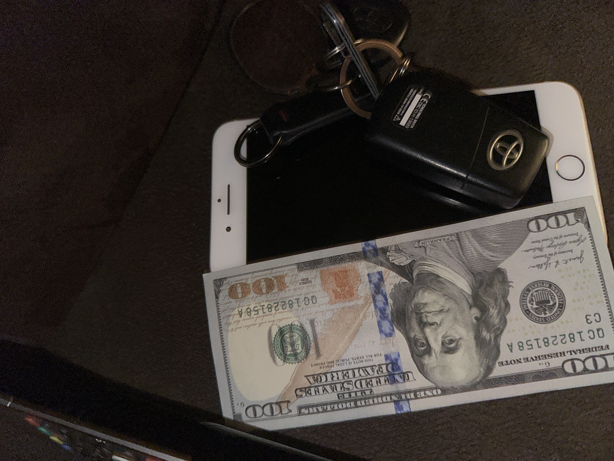 The image shows a white smartphone lying on a dark surface. A black car key with a key ring is on top of the phone. A one hundred dollar bill is partially covering the bottom portion of the phone. The objects are situated in a dimly lit environment. The image provides no indication of a location in Yerevan, Armenia.