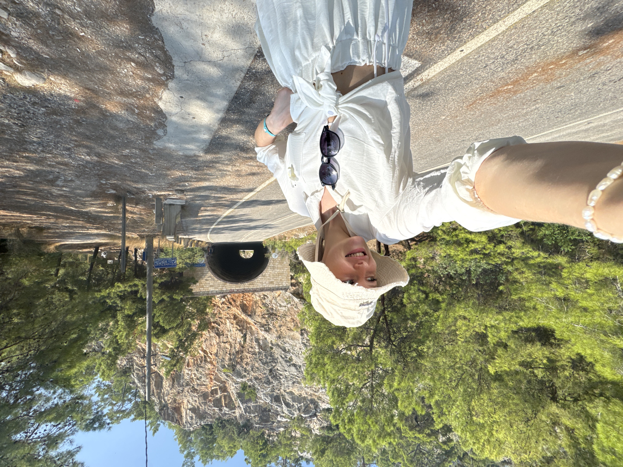 A young woman wearing a white bucket hat, white button-down shirt tied at the waist, and white skirt stands on an asphalt road, facing the camera and smiling. She has sunglasses hanging from her shirt. Behind her, a tunnel carved into a rocky mountainside is visible. The area is surrounded by green trees. A small pearl bracelet is on her left wrist and a blue bracelet is on her right wrist. This appears to be the Göynük Canyon entrance, a natural attraction near Kemer, Turkey, known for its hiking trails and canyon exploration.