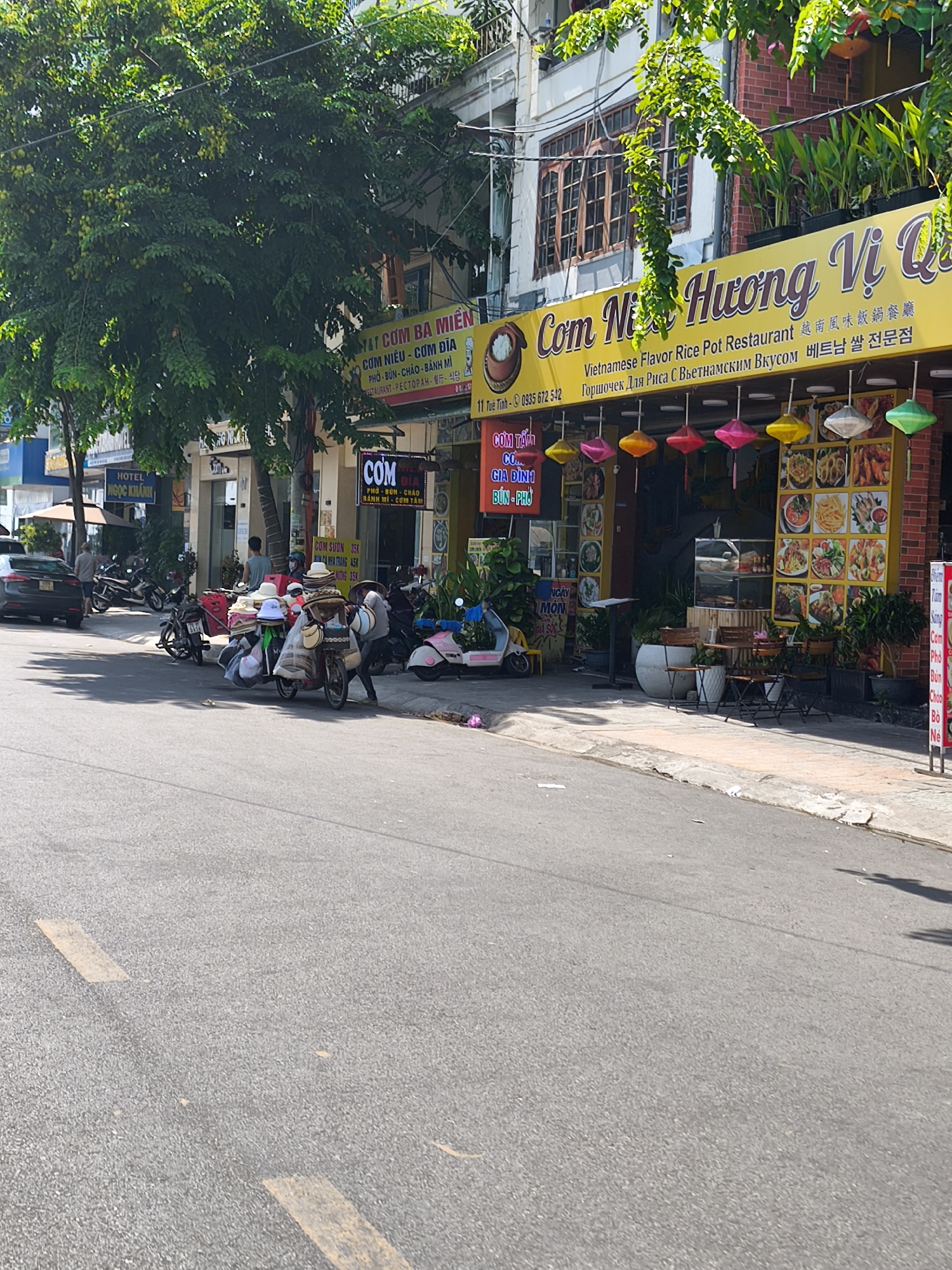 In Nha Trang Ward, Vietnam, a street scene unfolds with a focus on a restaurant and its surroundings. A brightly lit yellow sign, advertising "Com Ni Huong Vi Qu" and "Vietnamese Flavor Rice Pot Restaurant," dominates the upper portion of the image. Below this, a variety of colorful lanterns hang, illuminating a display of food pictures. In front of the restaurant, several tables and chairs are set up, suggesting an outdoor dining area. To the left, a parked car is visible, and further down the street, a row of motorcycles is lined up. A street vendor's cart, laden with a substantial collection of hats, is parked on the right side of the street, with a person tending to it. The overall environment depicts a busy commercial street in the city, with a mix of businesses and street life.