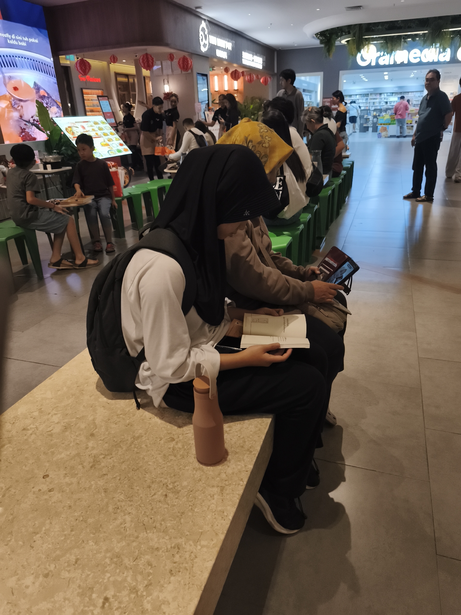 In a brightly lit indoor commercial space, likely a food court or shopping mall in North Jakarta, Indonesia, two individuals are seated on a beige bench. The person in the foreground, wearing a black hijab and a white long-sleeved shirt, is looking down at an open book in their lap. To their right, another individual, partially obscured by the first, is holding a tablet device in a burgundy case, also looking at the screen. A light brown bottle stands on the bench to the left of the book.
In the background, several other people are visible, some seated on green benches and others standing. One man in a dark blue shirt and black pants stands facing away from the camera. Other individuals are seen in the distance, engaged in various activities such as eating or shopping. The environment features a mix of retail displays, including a large digital screen advertising food and a sign that reads "Gramedia," suggesting a bookstore or media outlet. The floor is tiled, and hanging red lanterns add a decorative touch to the ceiling area. The lighting is a mix of overhead artificial lights and ambient light from the surrounding shops.