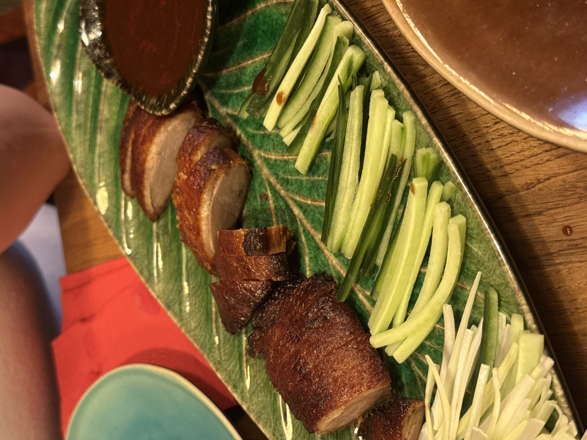The image depicts a plate of Peking duck with accompanying elements. Slices of roasted duck are arranged along the left side of a green leaf-shaped plate, beside a small bowl of dark sauce. To the right of the duck are lengthwise slices of cucumber and, further right, shredded scallions. The plate rests on a wooden surface and is surrounded by other objects including what appears to be a red napkin, a turquoise plate, and the edge of another plate containing a brown liquid. The location is assumed to be a restaurant in Sofia, Bulgaria.