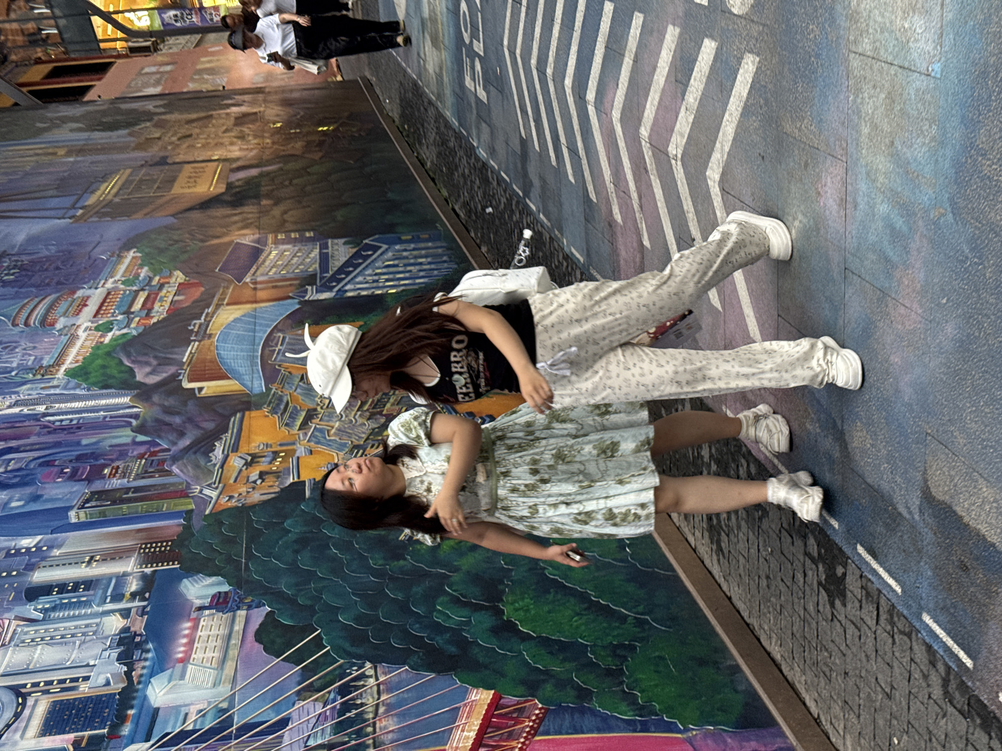 Two young women are walking along a sidewalk in Yuzhong District, China, with a vibrant mural of a city landscape on the wall beside them. The woman on the left wears a light blue floral dress and white sneakers, and her hair is dark. The woman on the right has on a white baseball cap, a black t-shirt with white text, and patterned light-colored pants, also wearing white sneakers and a white backpack. The mural features tall buildings, mountains, and a bridge, rendered in bright, varied colors. The sidewalk is paved with stone tiles and decorated with white painted arrows, indicating a pedestrian path. Further down the street, other pedestrians are visible in the distance.