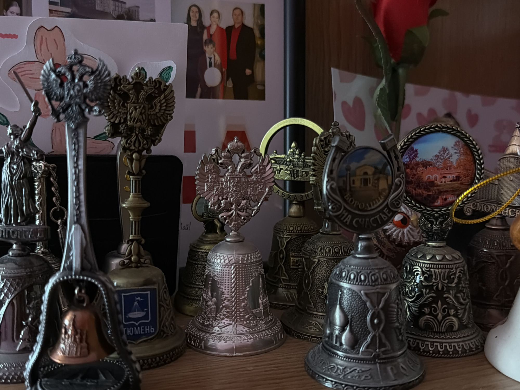 The image displays a detailed collection of decorative metallic bells, predominantly silver and bronze-toned, arranged on a wooden surface, likely a shelf, against a light background. These bells vary in size and design, with many prominently featuring the double-headed eagle, a national symbol of Russia.
Several bells are souvenirs, bearing inscriptions and imagery. One clearly visible bell is inscribed with "ТЮМЕНЬ" (Tyumen) and depicts a sailing ship, part of the city's coat of arms. Another bell reads "БОРОДИНО НА СЧАСТЬЕ" (Borodino for good luck) alongside an image of a building. Further inscriptions include "АБХАЗИЯ" (Abkhazia) and "СМОЛЕНСК" (Smolensk), which is partially visible on another bell. The word "ВОРОТЩИ" is also discernible on one bell. Architectural motifs, ornate patterns, and miniature depictions of buildings are common adornments across the collection. A unique bell is framed by a decorative spoon-like structure, containing a small bronze bell inside.
Behind the bell collection, a framed family photograph shows two adults and two children posing; the adults and a boy are smiling and dressed in festive attire, suggesting a celebration or special occasion. A red artificial rose stands among the items, and various paper items, including drawings and cards with heart patterns, are visible in the background.
The overall scene is an indoor still life, showcasing a personal collection of memorabilia. There is no direct activity taking place. The strong presence of Russian symbols and geographical references on the bells, such as Tyumen, Borodino, and Smolensk, reinforces the cultural context of Russia, consistent with the specified location of Perm, Russia.