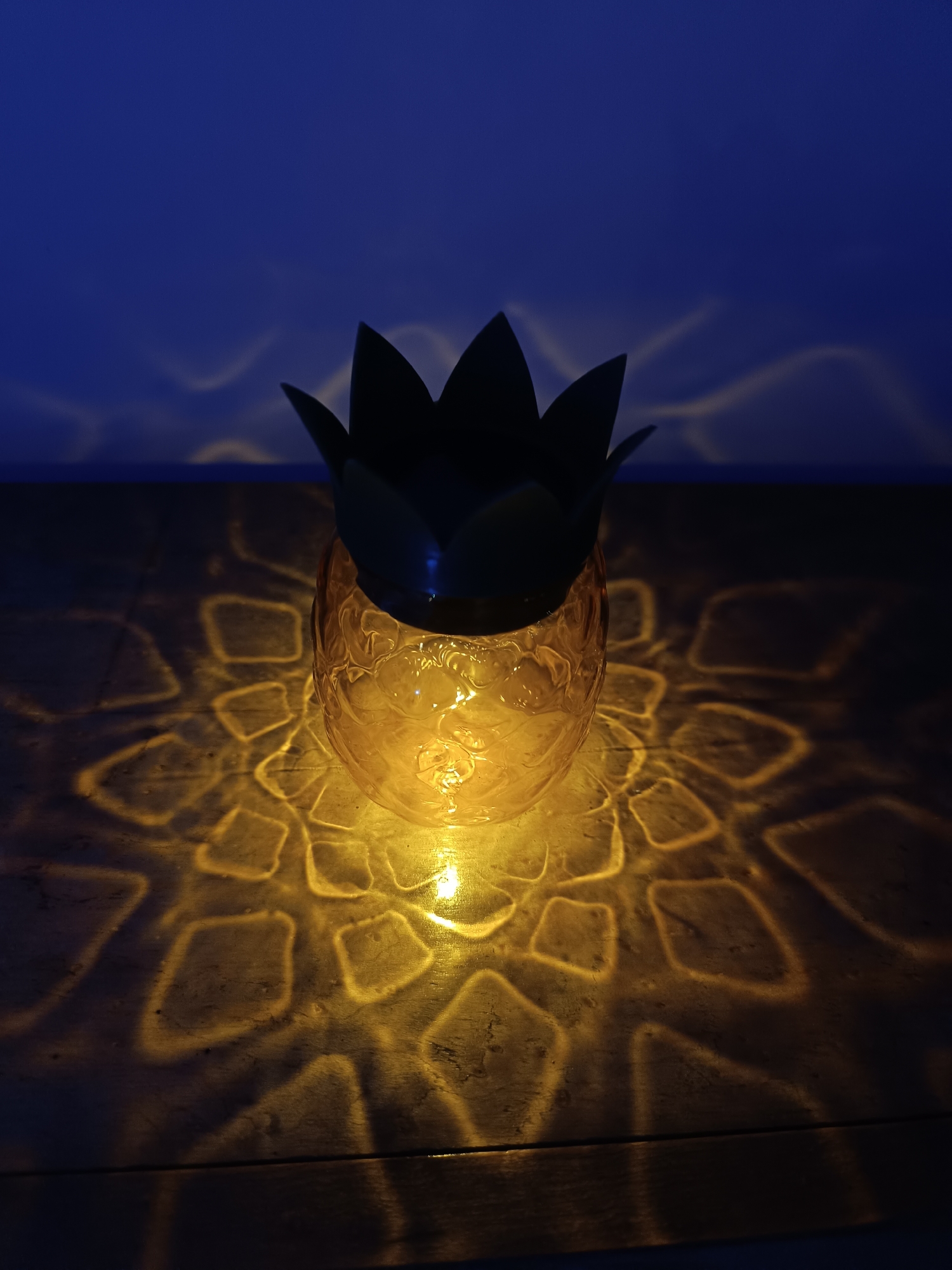 A pineapple-shaped lantern sits on a wooden surface, casting a patterned light on the table and wall behind it. The light is golden, and the room is dark, creating a dramatic contrast. The pineapple's "leaves" are silhouetted against the dark blue background. The origin or location of this image in Cherkovitsa, Bulgaria, is unknown.