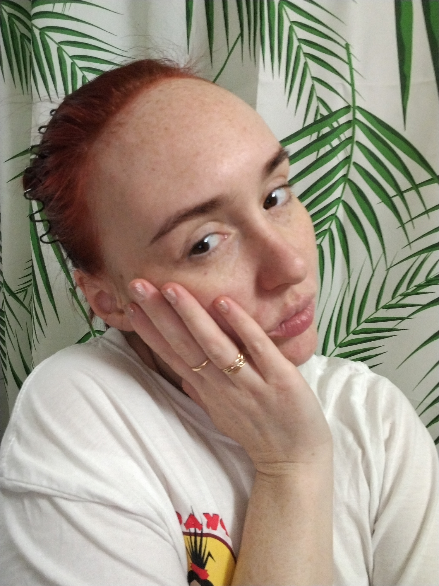 A light-skinned person with red hair, freckles, and a gold ring on their hand poses for a selfie. They are wearing a white T-shirt and are positioned in front of a patterned wall or curtain depicting green palm leaves. The person has their left hand touching their face and is looking at the camera. The location cannot be ascertained from the image provided.
