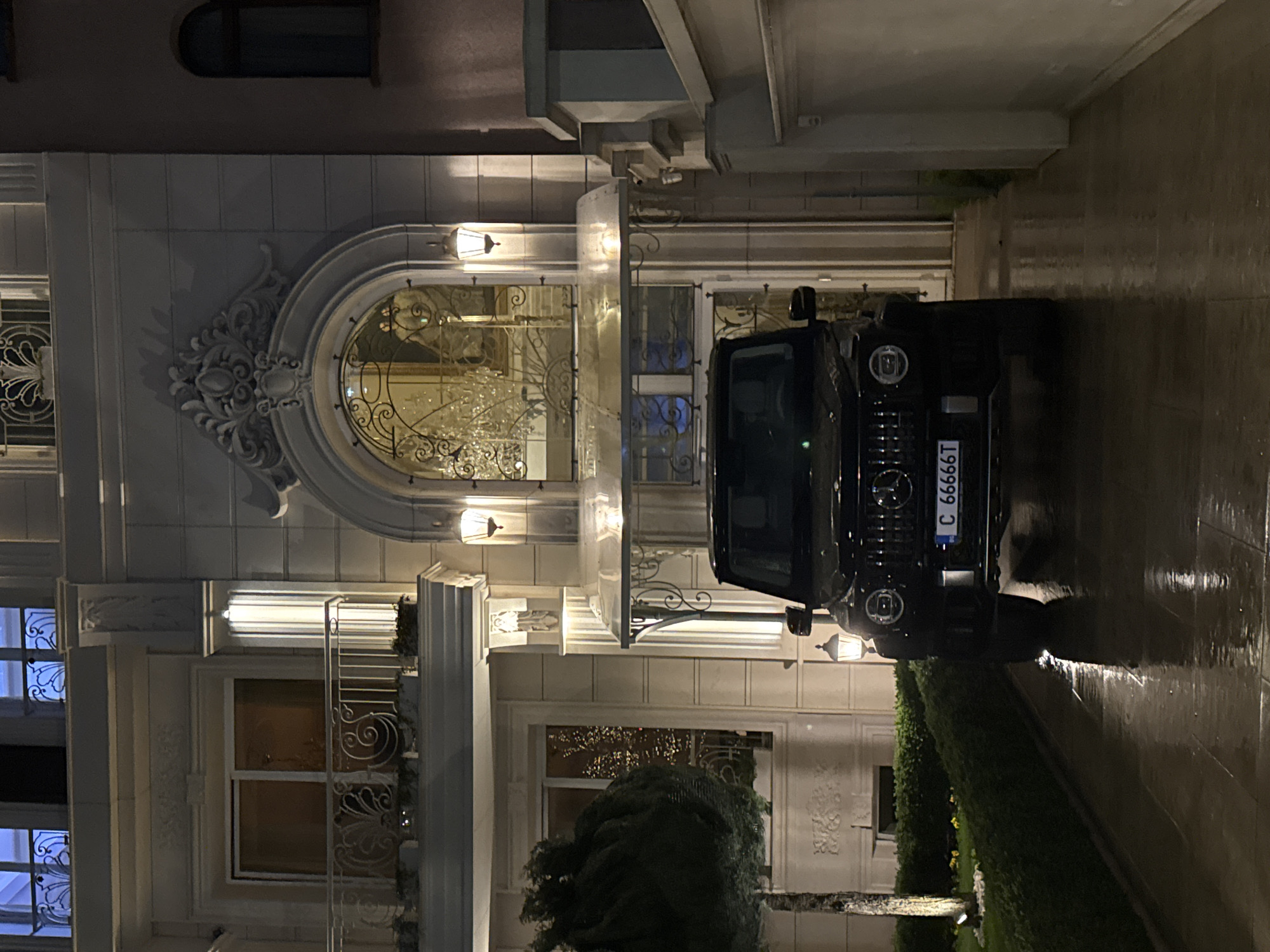 A dark-colored SUV is parked in front of a large, ornate building. The SUV has a license plate that reads "C 6666T". The building has multiple windows, a decorative archway, and ornate carvings on the facade. The environment appears to be outdoors at night, with wet pavement. The location could be the city of Sofia, Bulgaria.