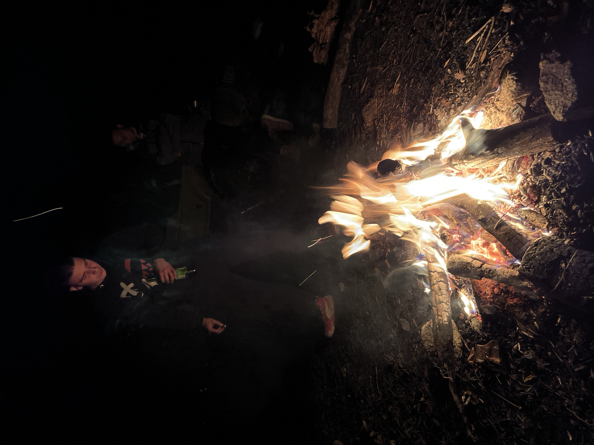A nighttime scene shows a burning campfire surrounded by dark earth and twigs. A person is visible to the left of the fire, appearing to hold a bottle and smoking a cigarette. Another person is partially visible in the background. The location is likely outdoors in a rural or undeveloped area near the city of Karavelovo, Bulgaria, as specified in the query.
