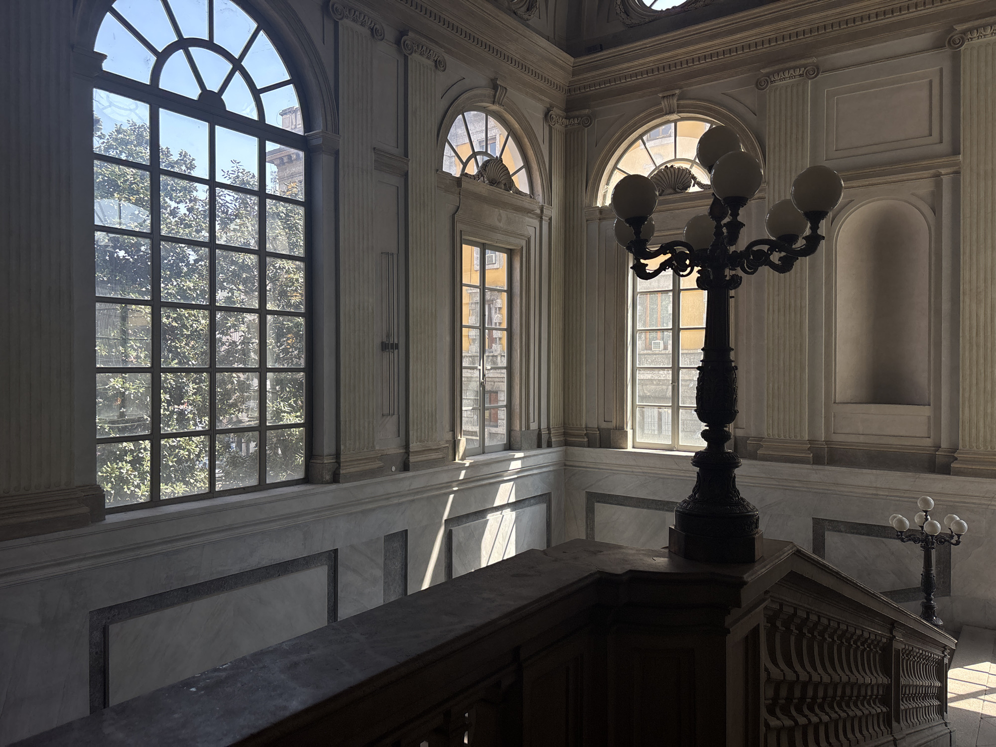 The image shows an interior view of a building in Milan, Italy. The building is characterized by a marble staircase with a dark wooden railing and ornate architecture with large arched windows. Light streams through the windows illuminating a portion of the marble wall. A dark, ornate candelabra stands atop the staircase landing. Another candelabra stands in the background along the wall. Outside the window, trees and buildings are visible. There are no people visible in the scene.