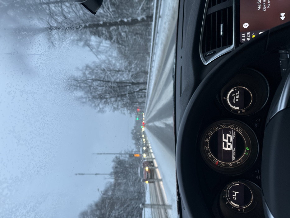 The image provides a forward view from the driver's perspective inside a vehicle on a snow-covered roadway in Lesnoy Gorodok, Russia. The windshield is partially obscured by a pattern of frozen precipitation. The multi-lane road ahead exhibits partially cleared asphalt with visible tire tracks, flanked by significant snow accumulation on the shoulders and a central median barrier. Bare, snow-laden trees line both sides of the road. Several vehicles with illuminated headlights and taillights are visible in the distance, traveling in both directions. Distant traffic signals display both red and green lights. The vehicle's dashboard is prominent in the foreground. The digital instrument cluster displays a current speed of 65 km/h, an odometer reading of 109,382 km, and a trip meter of 792.8 km. The transmission is engaged in 'D4', and the low beam headlight indicator is illuminated. A remaining range of 56.0 km is also indicated. To the right of the dashboard, an infotainment screen displays the time as 15:58, "LTE" network status, and partially visible Russian text, likely indicating media information. No individuals are visible within the frame.