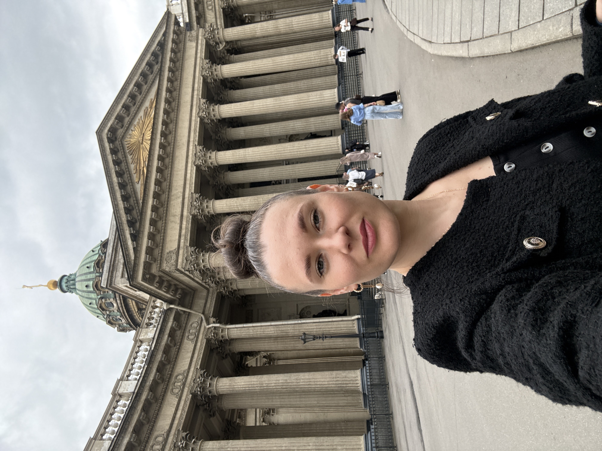 This is a selfie taken outdoors in front of the Kazan Cathedral in Saint Petersburg, Russia. A woman with her hair in a bun is in the foreground, wearing a black textured jacket over a black top. She has on dangling earrings. Behind her, the facade of the Kazan Cathedral is visible, with its semicircular colonnade and dome. Several other people are walking around in the plaza in front of the cathedral, and a lamppost is positioned between the woman and the building. The sky is overcast.
