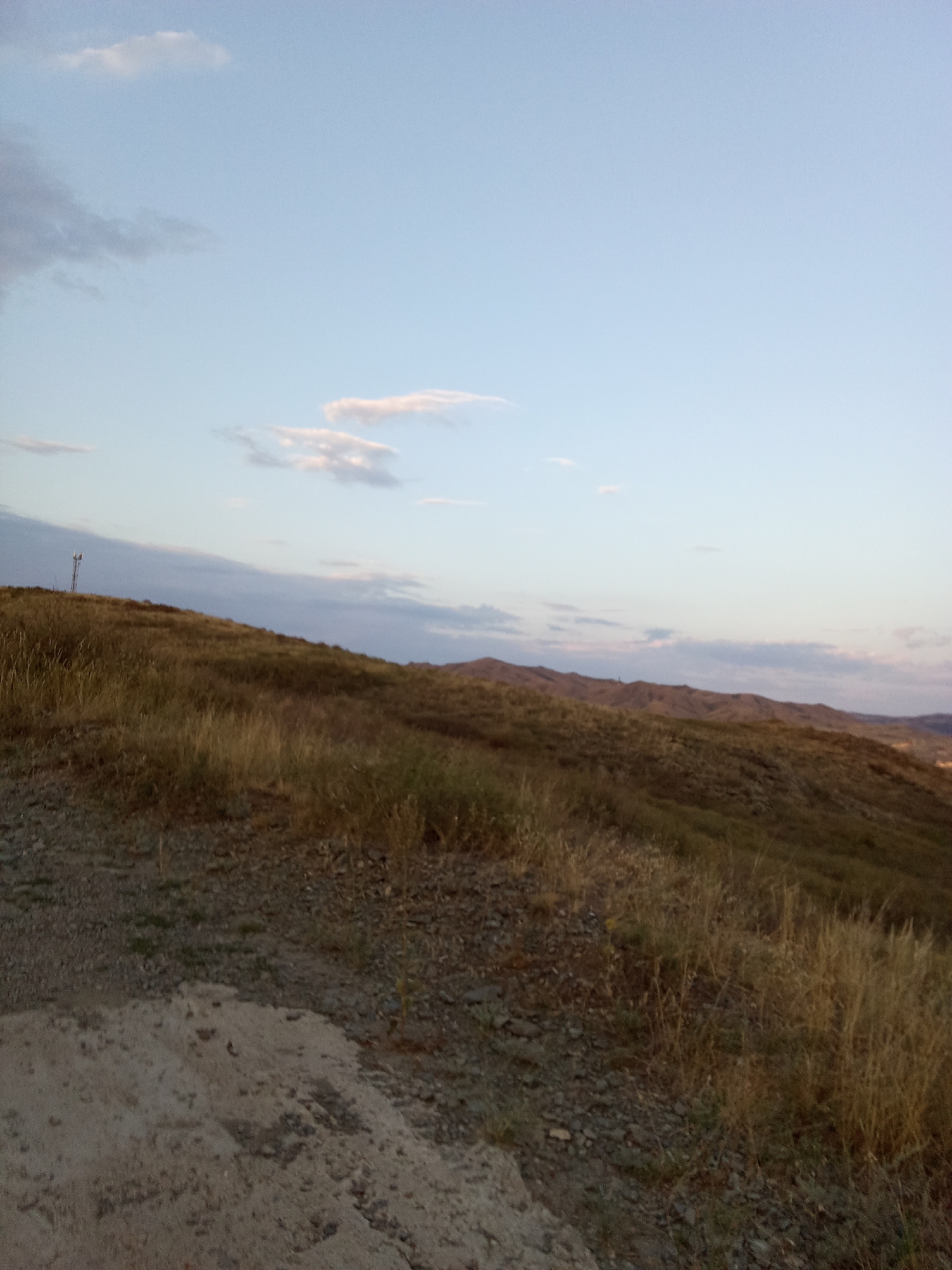 The image depicts a wide, grassy hillside under a pale blue sky with scattered wispy clouds. The terrain is uneven, with dry, golden grasses and scattered rocks covering the slopes. In the foreground, a patch of lighter-colored earth, possibly sand or gravel, is visible. Further back, rolling hills and distant mountains form the horizon. A single, tall tower, likely a communication mast, stands on the crest of a hill to the left of the frame. The scene appears to be an outdoor, natural landscape, possibly on the outskirts of a rural or semi-rural area.