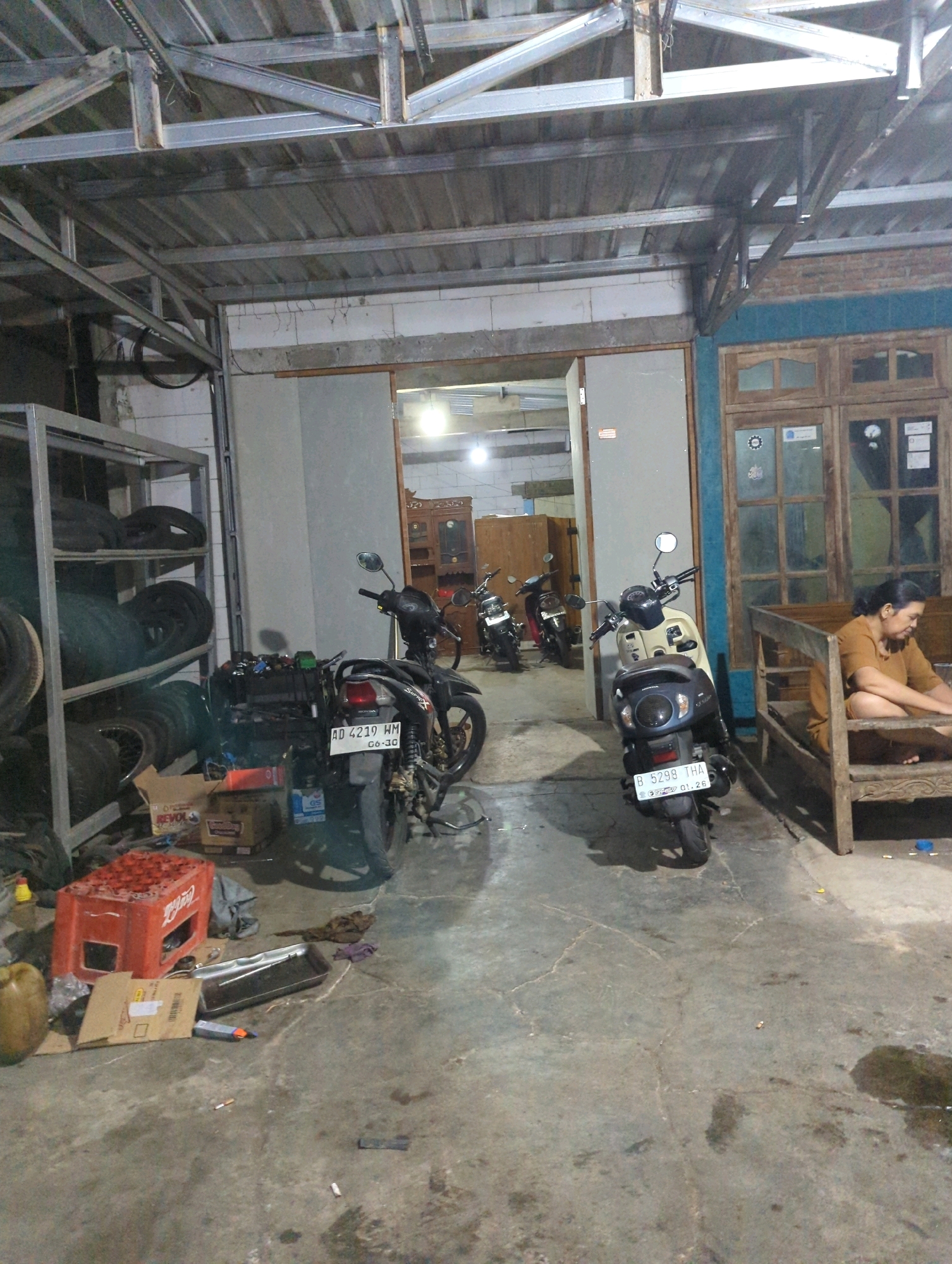 The image depicts an outdoor covered area, likely a workshop or storage space, in Senggrong, Indonesia. The scene is cluttered with various objects, including several motorcycles parked haphazardly. In the foreground, a dark-colored motorcycle with a license plate "AD 4219 WM" is positioned. To its right, a lighter-colored scooter with a license plate "B 5299 THA" is also present. To the right of the motorcycles, a woman wearing an orange shirt is seated on a wooden bench. The environment appears to be a concrete floor with some debris and puddles of liquid. A metal shelving unit filled with tires is visible on the left side of the image, along with a red Coca-Cola crate. In the background, there are doorways leading to interior spaces with wooden furniture and windows. The overall impression is one of a functional, yet somewhat unkempt, utilitarian space.