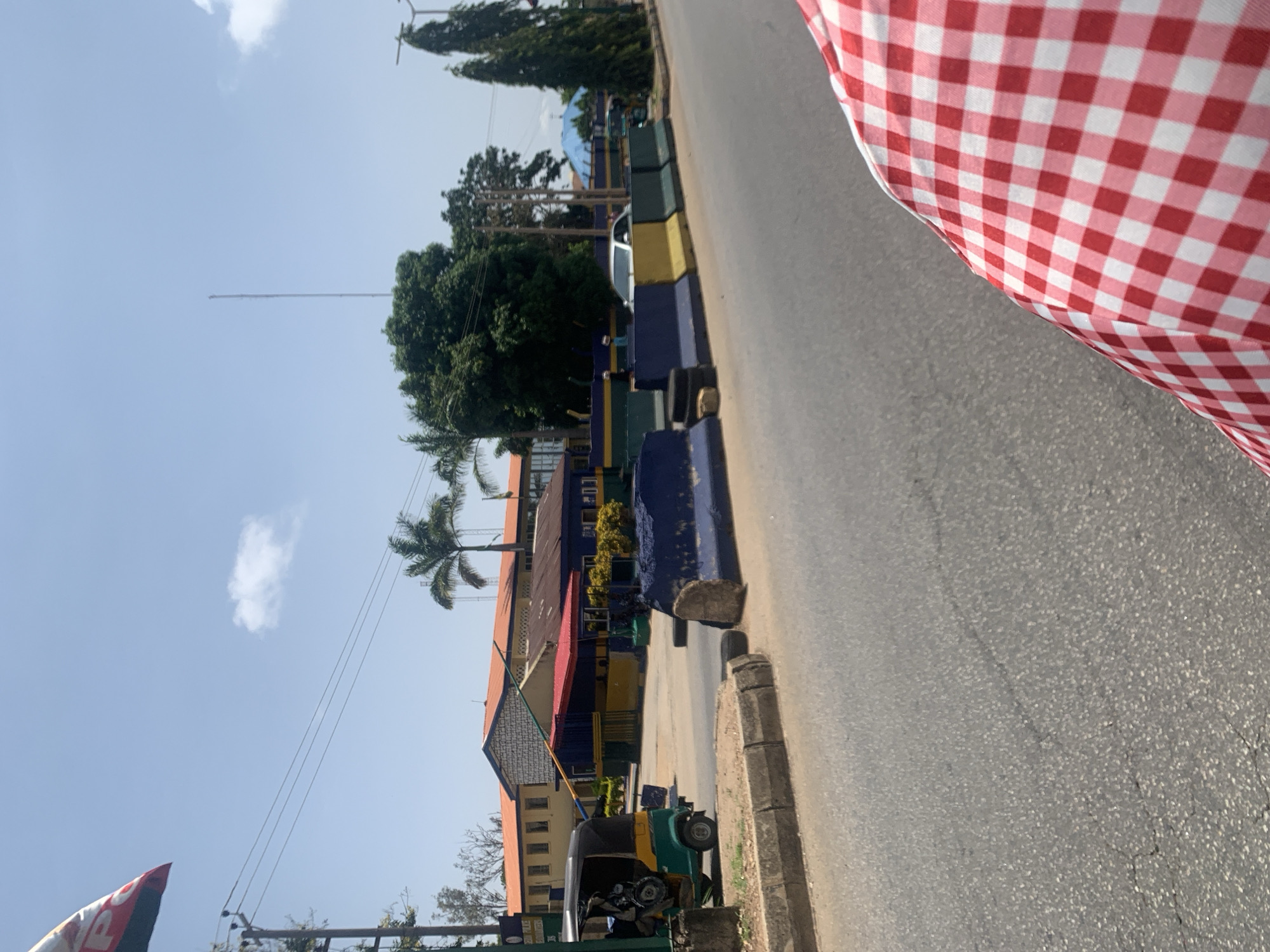 The image captures a scene in Jos, Nigeria, with a road in the foreground and a building complex in the background. The road appears to be asphalt, showing some signs of wear. On the right side of the road, there are concrete barriers painted in blue, yellow, and green, with some tire remnants. A white sedan is visible behind the barriers, and a tuk-tuk, a three-wheeled motorized vehicle, is parked to the left of the road, near a building entrance.
The building complex features orange-tiled roofs and yellow and blue painted walls, possibly indicative of a government or institutional structure. Green foliage, including palm trees and other trees, surrounds the building and extends into the background. A tall, thin antenna or tower is visible in the distance. The sky is clear blue with a few scattered white clouds, suggesting a sunny day.
In the lower right corner of the image, a close-up view of a red and white checkered fabric is prominent, suggesting the photo might have been taken from a vehicle with a patterned interior or from someone holding such fabric. A flag with red, white, and blue colors, possibly with lettering, is partially visible in the upper left corner. The overall impression is an urban street scene in a location that appears to be experiencing warm weather.