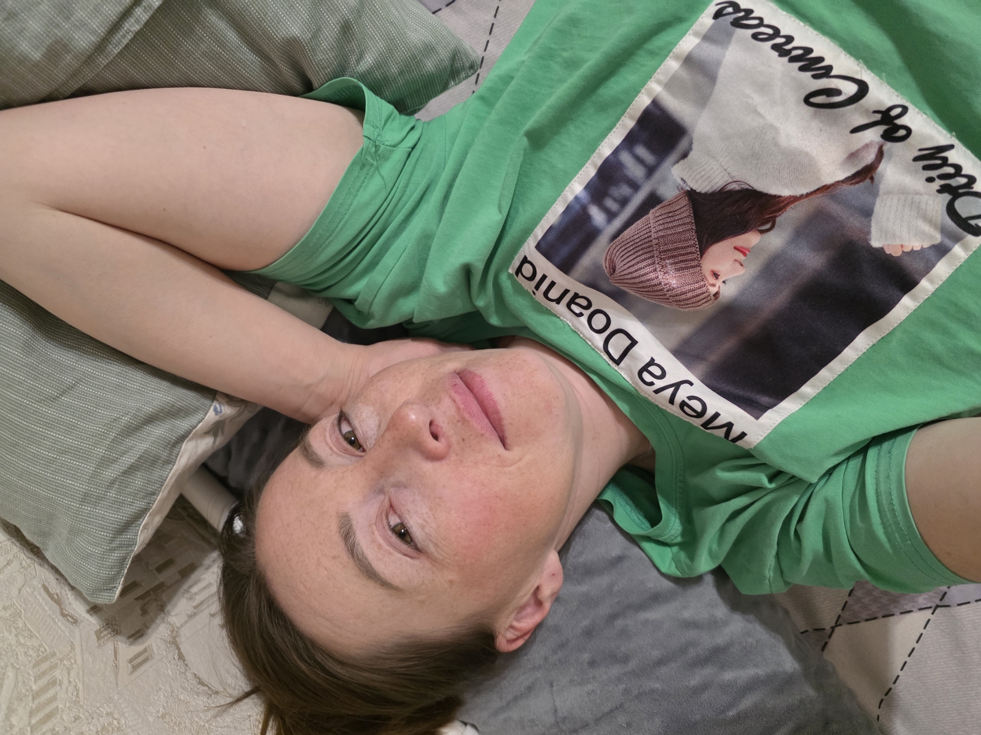 A woman is lying down on a bed. She is wearing a green t-shirt with a square design on it that contains a picture of a person wearing a hat and the text "Meya Doania" and "City of Caneas". The bed is covered with pillows and a patterned blanket. Her location in Chaikovsky, Russia cannot be determined from the image.