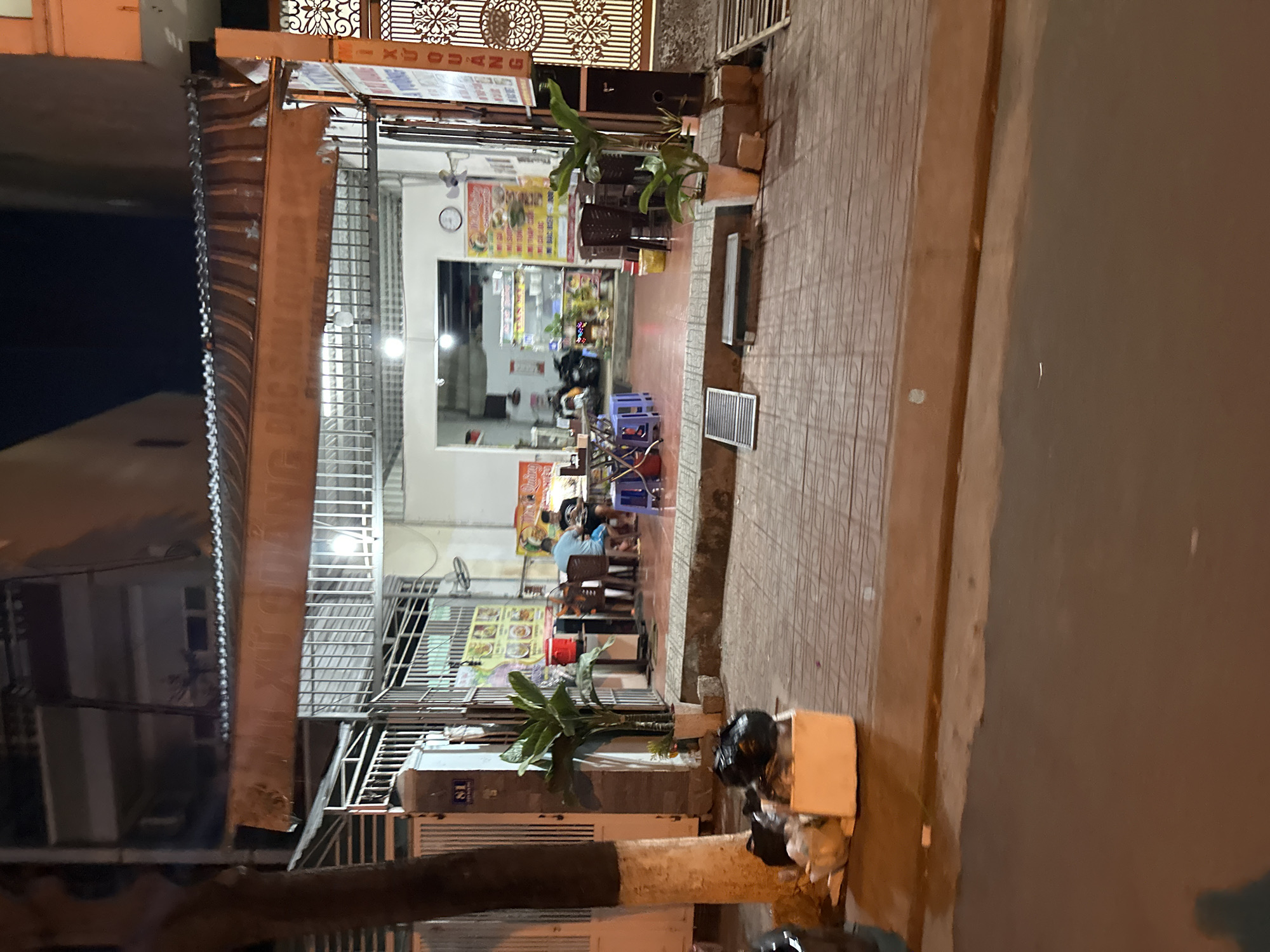 The image depicts a street scene in Bac Nha Trang Ward, Vietnam. The main focus is a restaurant or eatery, open to the street, with several patrons seated at tables, presumably eating. The establishment is lit with overhead lights. Outside, the pavement leads to a road, and a motorcycle is parked nearby. Plants are placed outside the establishment. A street address or number is visible. The overall impression is of a relaxed, informal atmosphere.