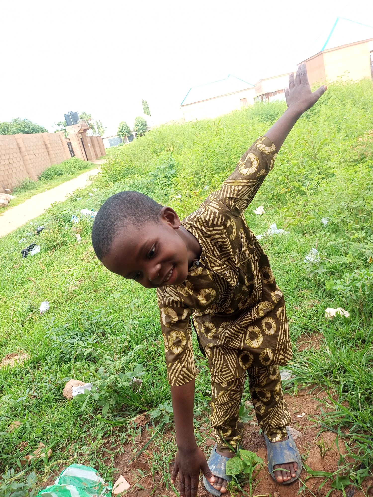 In the city of Tempure, Nigeria, a young boy is captured outdoors, leaning forward and smiling at the camera. He is dressed in a brown and gold patterned outfit, consisting of a tunic and pants, and wears blue sandals. His right arm is raised. The background is a grassy area with some scattered debris. Buildings and a brick wall are visible in the distance, suggesting a semi-urban or rural setting. The sky is bright and overcast.