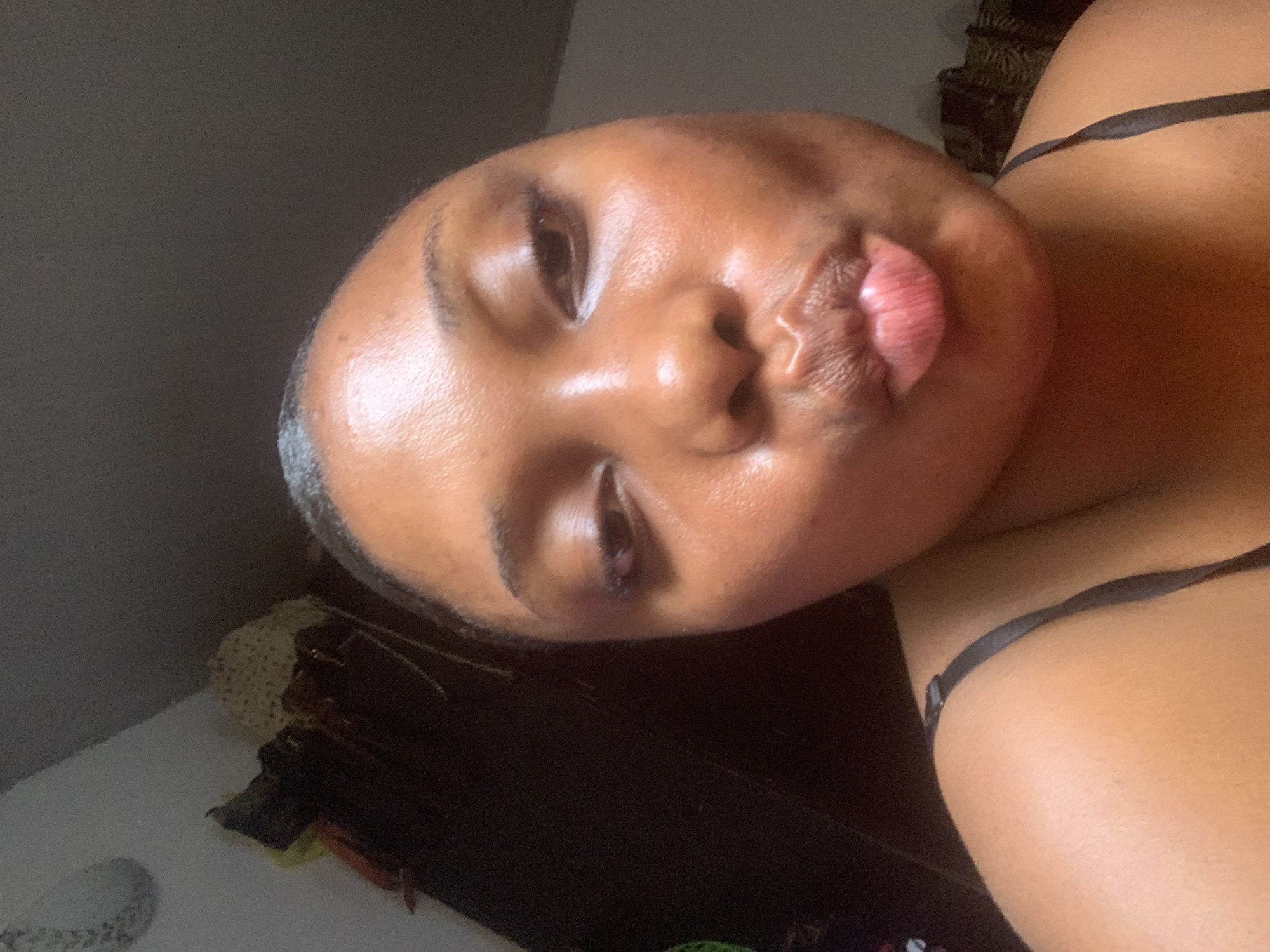 A close-up, portrait-style photograph depicts a person with brown skin making a pouting expression, commonly known as "duck face." The person's eyes are partially closed, and the lips are prominently puckered. They have short hair. A black strap, possibly from a garment, is visible. The background is dark and indistinct, but some items are discernable, including a white wall and brown wooden objects. The location in Abuja, Nigeria, is unknown and cannot be identified based on the image alone.