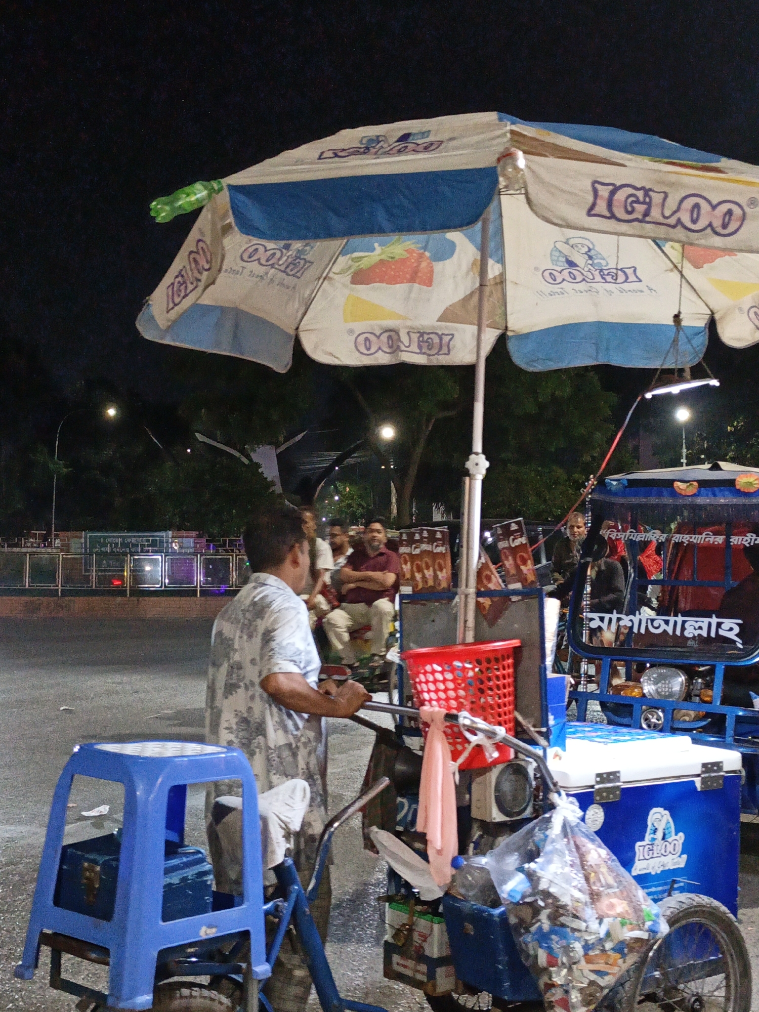 An outdoor, nighttime scene depicts an ice cream cart with a blue and white Igloo umbrella. A person wearing a light-colored floral shirt stands behind the cart, holding its handle. To the right, a blue motorized rickshaw is parked. Several people are seated in the rickshaw, and text in an Arabic script is visible on its side. The background shows trees and streetlights. The ground is paved.