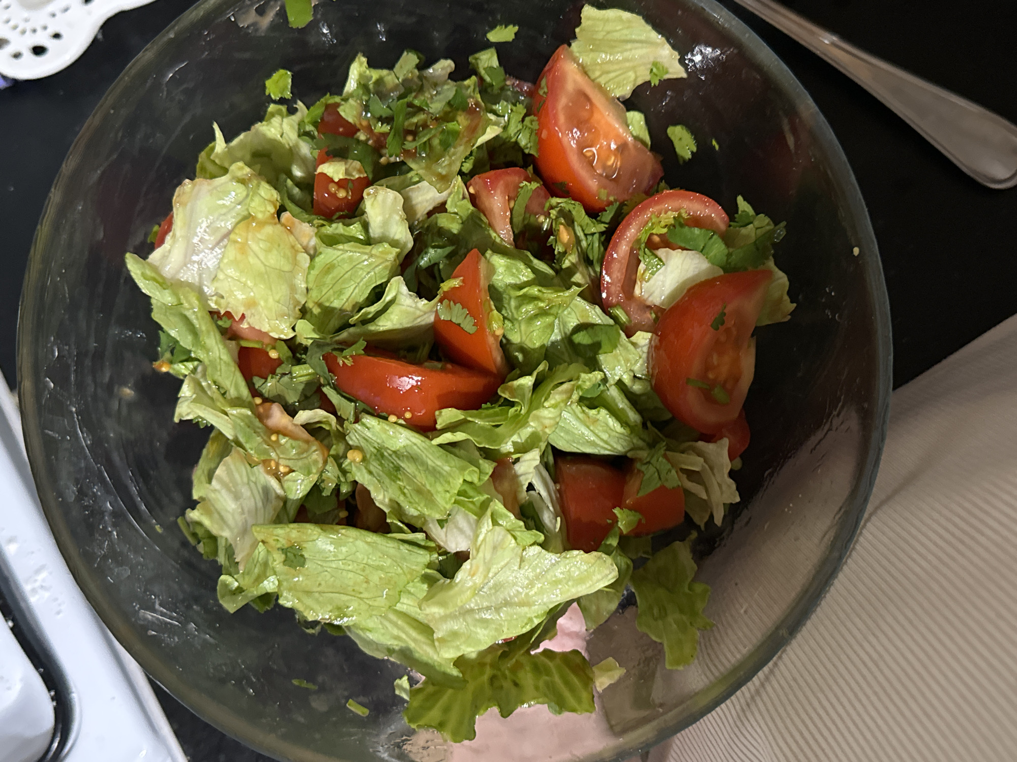 A vibrant salad, served in a clear glass bowl, is the central subject of the image. The salad primarily consists of fresh green lettuce leaves and wedges of ripe red tomatoes. Interspersed among the lettuce and tomatoes are finely chopped green herbs, likely cilantro or parsley, adding a contrasting touch of color. A glistening dressing, possibly an oil and vinegar mixture, coats the salad components, suggesting it has been freshly tossed.
The bowl rests on a dark, reflective surface, which appears to be a table or countertop. To the top left, a white, intricately designed object with cut-out patterns is partially visible, possibly a decorative placemat or coaster. On the right side of the main image, a metallic eating utensil, likely a fork or spoon, lies on the dark surface beside the bowl. Another crop reveals a light-colored, possibly striped, fabric surface extending from the bottom right of the bowl, suggesting a napkin or tablecloth. The lighting is even, implying an indoor setting, possibly a kitchen or dining area. There are no people visible, and no discernible text. The scene depicts a prepared dish ready for consumption.