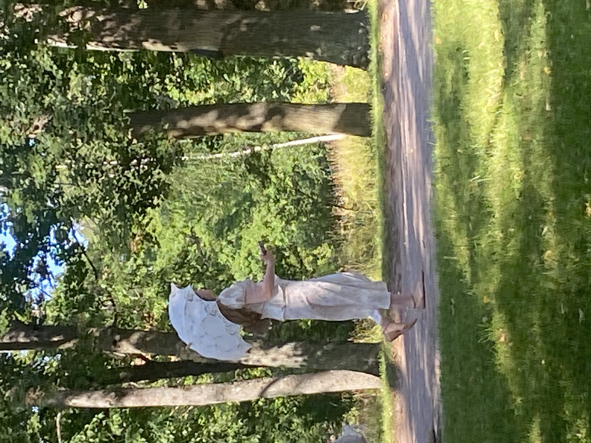A person in light-colored, loose-fitting clothing and sandals walks on a paved path in a park. The person holds a white, decorative umbrella over their head and carries a small, dark object in their right hand. The path is bordered by green grass and surrounded by trees with dappled sunlight filtering through the leaves. The scene appears to be set in a natural environment, possibly during daytime.