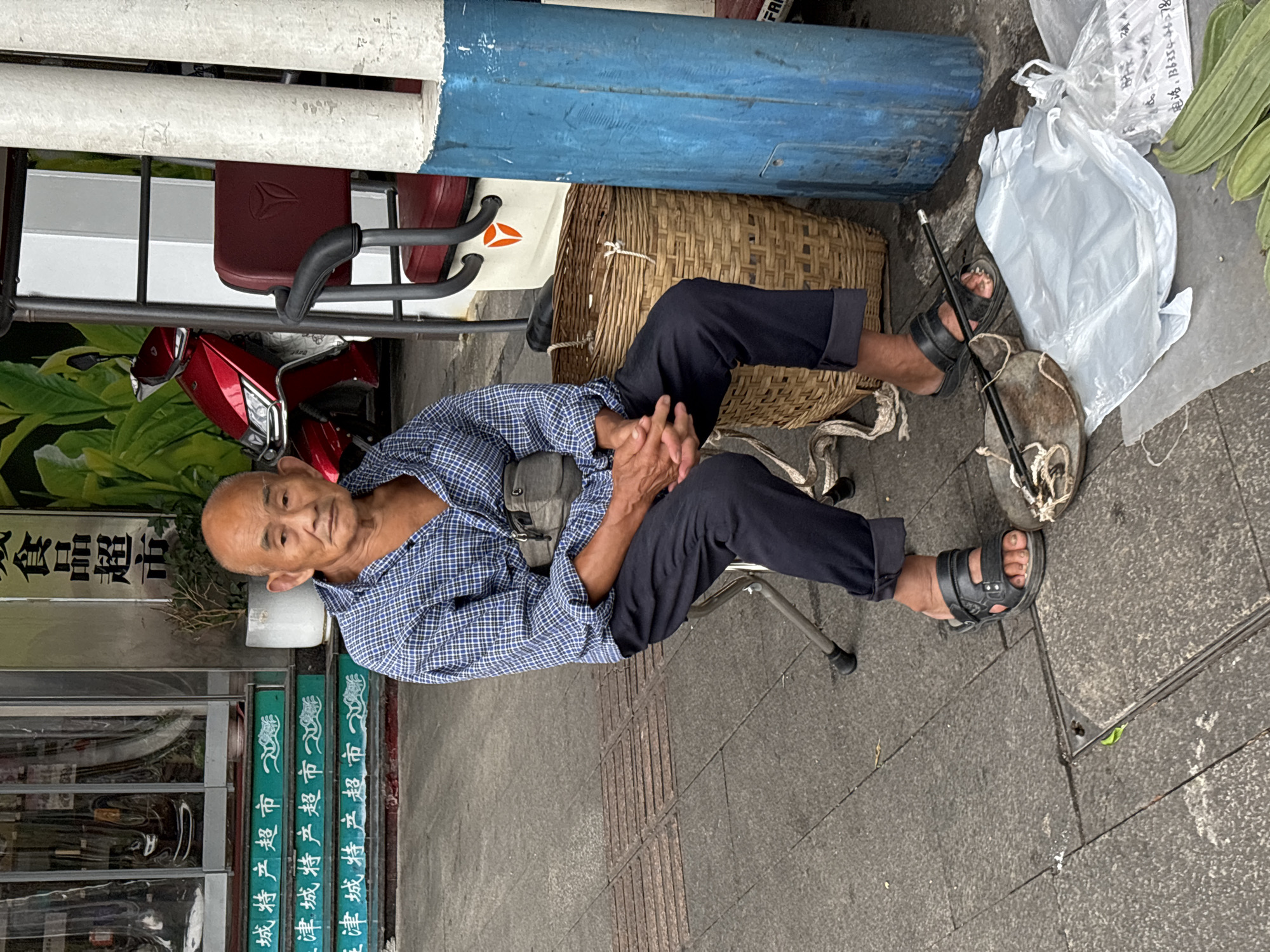 An elderly East Asian man with a bald head and wearing a blue checkered shirt and dark pants sits on a small metal stool outdoors. He has a gray fanny pack around his waist. In front of him, on the ground, is a small weighing scale with a long handle and a white plastic bag. To his right, there is a large woven basket and a blue cylindrical object. Behind him, a red scooter is visible, as well as the entrance to a store with green steps that have Chinese characters on them. The scene appears to be on a sidewalk. The store's signage and the characters on the steps suggest the location is Jiangjin, China.