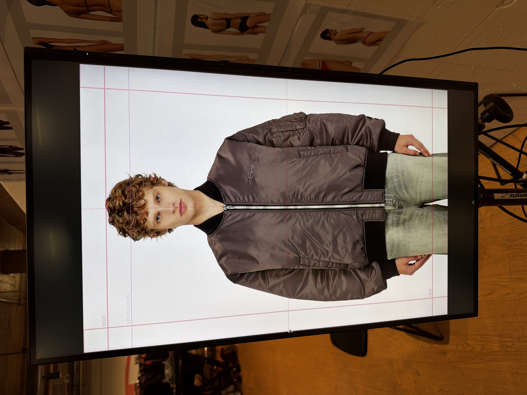 The image shows a flat-screen monitor displaying a studio shot of a person wearing a brown bomber jacket and jeans. The screen has faint vertical and horizontal lines visible, presumably guides used during the photo shoot. The monitor is set against a backdrop of what appears to be a room with promotional posters of people in swimwear. The environment includes a wooden floor and photographic equipment, suggesting a studio or exhibition setting. While it's impossible to independently verify the city of Mirabella, Italy, based on the image content alone, if the image was provided in that context, then the location is an exhibition or studio in Mirabella, Italy.