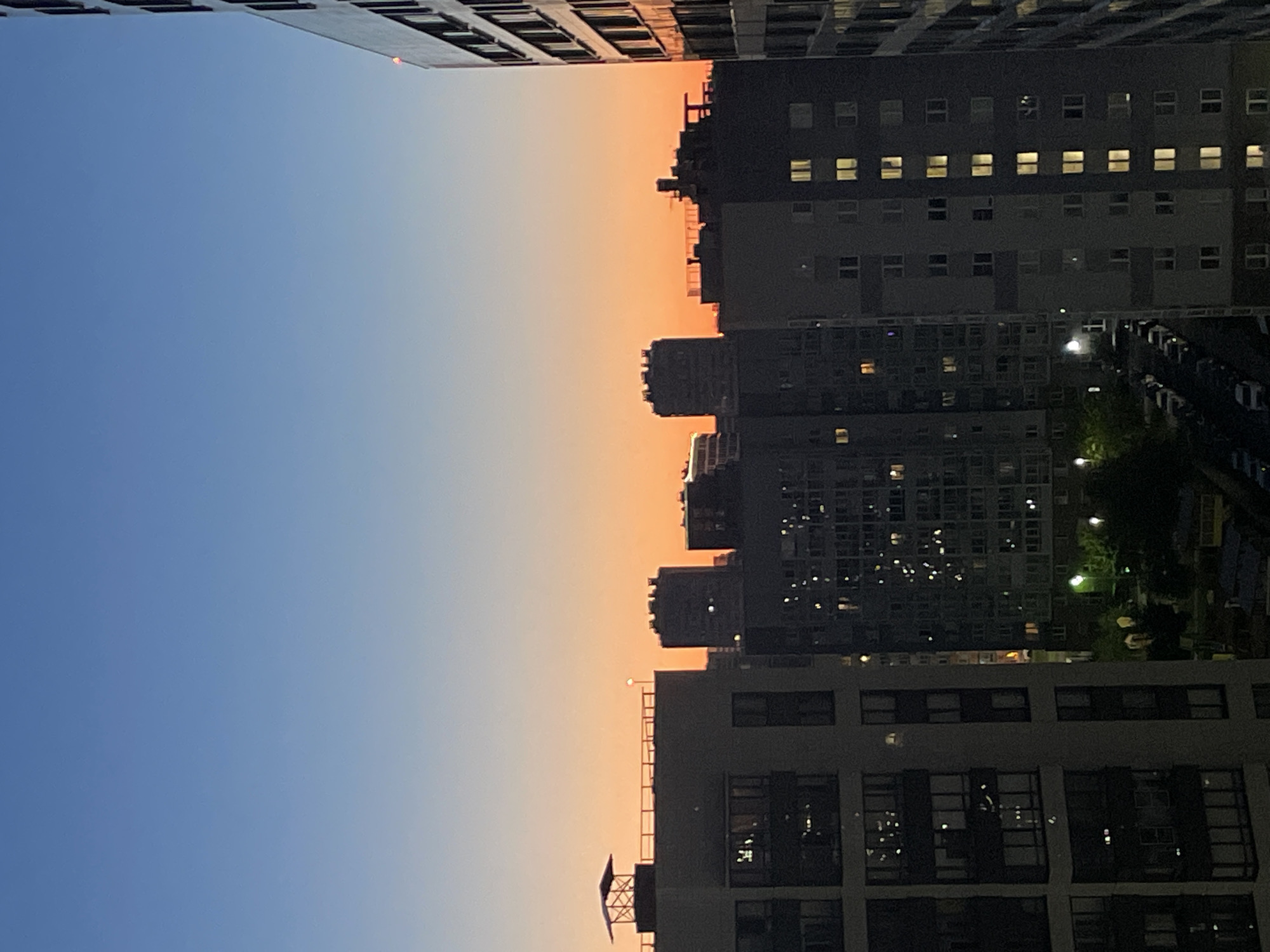 The image captures an urban cityscape at dusk, with the sky transitioning from a deep blue at the top to a soft orange horizon. Silhouetted against this sky are several multi-story apartment buildings. The building in the foreground on the left is partially cropped, revealing its facade with numerous windows. To its right, a cluster of darker buildings rises, their windows illuminated with warm, artificial light, suggesting occupancy. Further into the distance, more buildings of varying heights and architectural styles are visible, some with distinct shapes, like a building with a prominent, stepped roofline. Streetlights are visible between the buildings, casting a glow on the scene. The overall impression is of a residential area in a city during the evening twilight. The location is specified as Sinyavino, Russia, which is known for its residential development and proximity to St. Petersburg.