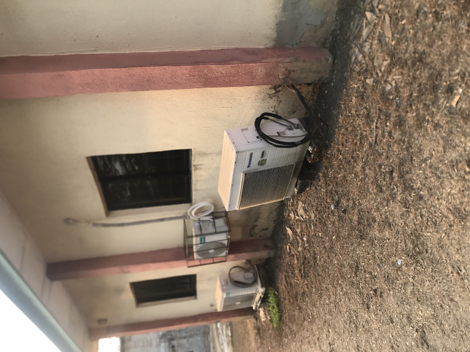The image displays an exterior view of a building wall and the dry ground beside it, set in Umudike, Nigeria. The building features a light-colored, possibly cream or beige, wall with a prominent reddish-brown horizontal beam or trim running across it. Two dark, rectangular openings, resembling windows or vents, are visible in the wall.
Lining the base of the wall, on the ground, are three outdoor air conditioning units. The most prominent unit, positioned towards the top right of the frame, is white and bears the brand name "Panasonic" clearly visible on its side. Black hoses or cables are coiled on top of this unit. Further down the wall, another air conditioning unit is partially enclosed within a black metal cage or frame, with white tubing or coils nearby. A third similar white unit is visible further along the wall towards the bottom of the frame. A grey electrical conduit or pipe runs along the wall, connected to one of the units.
The ground adjacent to the building is dry and covered in sparse, light-brown vegetation, appearing like dry grass or dirt, interspersed with small rocks and some scattered debris. The lighting suggests it is daytime, with clear weather conditions. There are no people visible, and no specific activity is taking place in the scene.