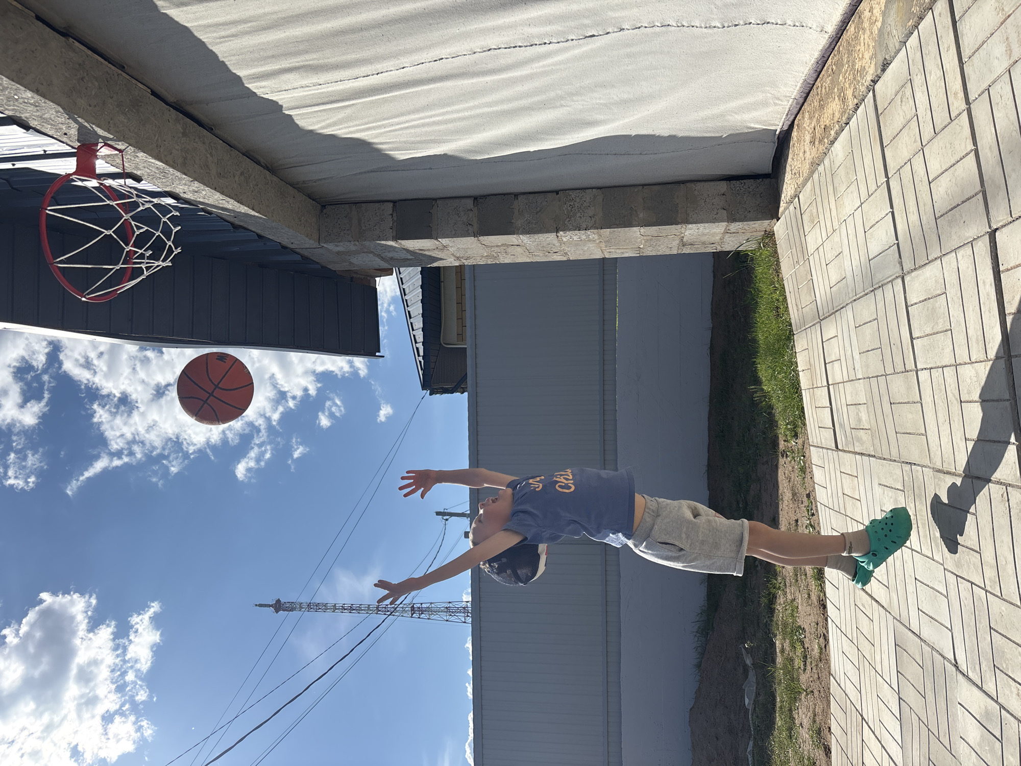 This outdoor photograph, taken in the city of Chaikovsky, Russia, features a young child captured in mid-air, having jumped towards a basketball. The child wears a blue t-shirt with orange text, grey shorts, grey socks, and teal clogs, with a dark baseball cap worn backwards. Both arms are extended high above the head, hands open, and the mouth is agape. An orange basketball is visible in the air, positioned slightly above and to the right of the child's hands, in line with a red basketball hoop equipped with a white net.
The basketball hoop is mounted on a light-colored, possibly concrete, structural component of a building on the right side of the frame. Above the hoop, the building's facade consists of dark grey corrugated metal siding. Below the hoop, there is a large, light-colored, textured vertical surface, possibly a rolled-up garage door or curtain. A masonry pillar forms the corner of this building section. The immediate foreground is paved with light grey rectangular stones arranged in a herringbone pattern. Beyond the paving, to the left and rear, are areas of exposed dirt and sparse green grass, bordered by a light grey corrugated metal fence.
The background features a bright blue sky with scattered white cumulus clouds. Power lines crisscross the upper left portion of the frame, and a tall, lattice-structured communication tower is prominently visible in the distance on the left. Strong sunlight from the left side of the frame casts distinct shadows of the child and other objects onto the paved ground.