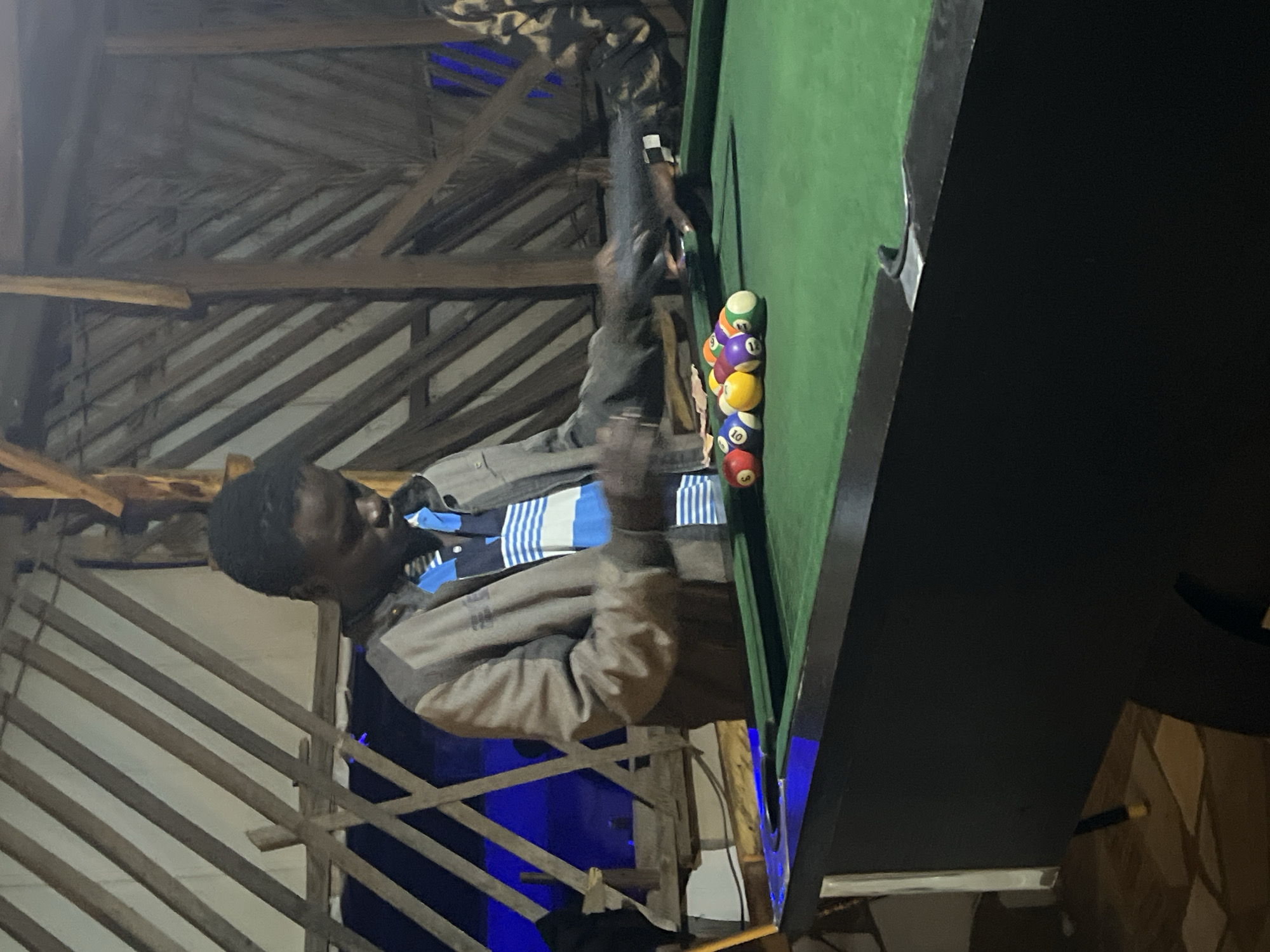 A young Black male, wearing a grey jacket over a blue and white striped shirt, stands over a green billiard table in what appears to be an outdoor or semi-outdoor establishment in Durumi, Nigeria. He is in the process of setting up billiard balls, which are clustered on the table in front of him, with some balls having visible numbers such as 3, 9, and 10. The background shows a rustic, slatted wooden structure, possibly a roof or wall, with some bamboo or palm frond elements visible. Blue lights are visible in the background, suggesting ambient lighting. The overall impression is of a casual recreational setting.