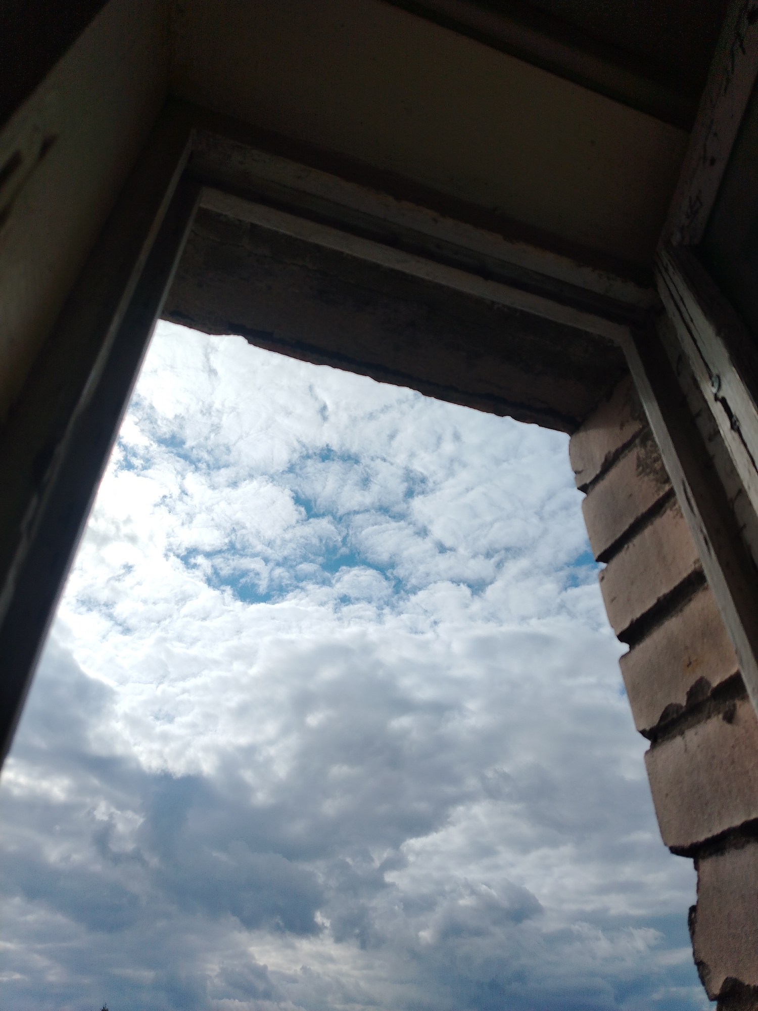 The image is a low-angle, wide shot looking up through a rough, unfinished doorway or window frame towards a cloudy sky. The frame is constructed of dark, weathered wood on the left and top, and roughly laid bricks on the right. The sky is filled with a mix of fluffy white and darker, heavier grey clouds, with patches of blue sky visible in between. The overall scene suggests an abandoned or under-construction building, possibly in an industrial or historical area. The specific location in Vyborg, Russia is not discernible from the visual information alone. There are no people or discernible actions taking place.