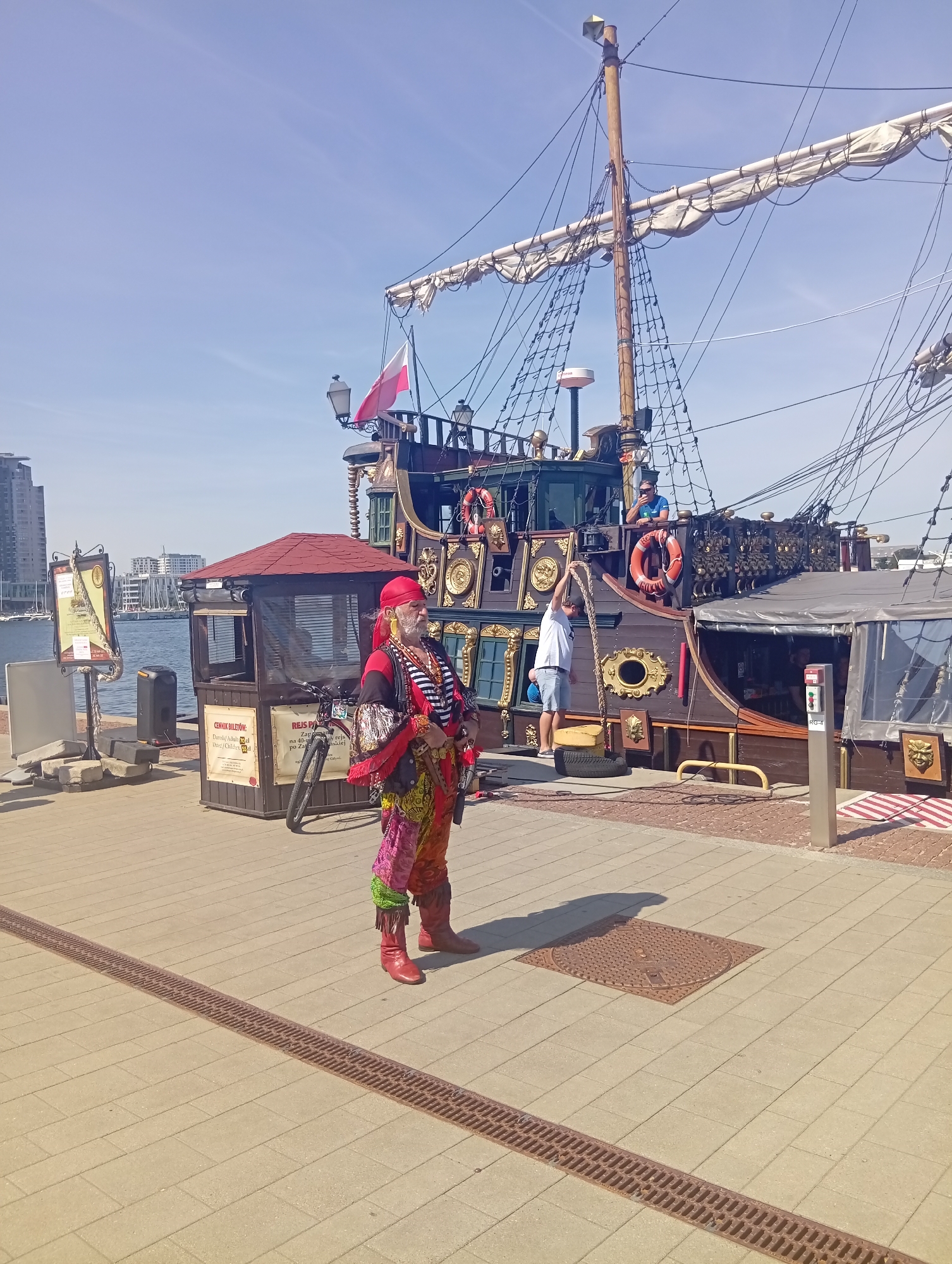 A person dressed in a colorful, pirate-like costume stands on a paved promenade in Gdynia, Poland, with a tall-masted ship docked behind them. The person, who appears to be an entertainer, has a red bandana, a black and white striped shirt, and vibrantly patterned pants tucked into red boots. They are holding a rope in their right hand and have a beard and mustache. To the left of the person is a small ticket booth with a red roof and blinds. A bicycle is parked next to the booth. The ship, which has a Polish flag flying from its mast, is intricately decorated with gold accents and several windows. The promenade overlooks a body of water, with modern buildings visible in the background. The sky is clear and blue.