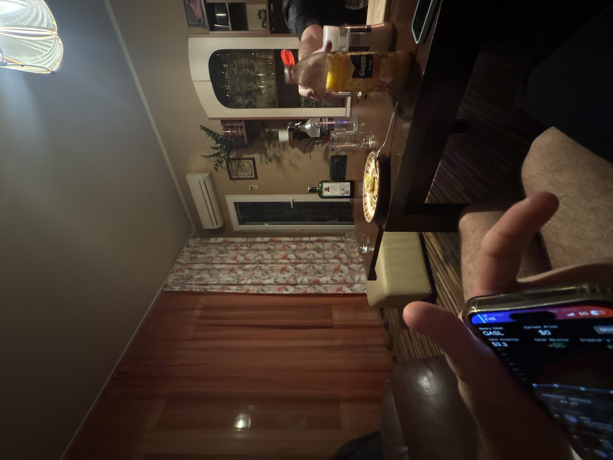 This image depicts an indoor scene, identified as a living room in Sofia, Bulgaria, observed during what appears to be an evening or nighttime social gathering.
Two individuals are partially visible within the frame. In the immediate foreground, a person's left hand holds a smartphone, displaying an application interface with text elements including "OASL" and numerical values. A portion of this individual's legs is also visible, indicating a seated position. On the right side of the dark wooden coffee table, another person's arm and hand, clad in dark attire, are partially visible as they reach towards the table's contents.
The central dark wooden coffee table holds several items: on the left, a decorative plate with a single light-colored food item and a fork; in the center, an empty clear drinking glass. A green bottle identified as Jägermeister is positioned behind the plate, and a bottle of Russian Standard vodka stands behind the glass. To the right, a plastic bottle of Cappy Pulpy orange juice is present alongside a white shaker or container. A second, lighter-colored smartphone is partially visible near the table's edge on the right.
The room's furnishings include a brown leather armchair or sofa, partially visible on the left, and a light beige ottoman situated to the right of the table. The walls are a light beige color, featuring white crown molding along the ceiling line. A large window on the left is covered by opaque reddish-brown sheer curtains, while an adjacent window features patterned curtains with white, red, and orange tones. A white wall-mounted air conditioning unit is fixed to the upper left wall. Decorative elements include framed pictures and potted plants. A white door is visible on the left wall, and a tall, white cabinet with an arched opening, displaying glassware, is prominent on the right wall, flanked by shelves holding various items. The floor is covered with a patterned wooden laminate or parquet. The ceiling is plain and light-colored, with a decorative light fixture visible in the upper right corner, emitting a warm glow.