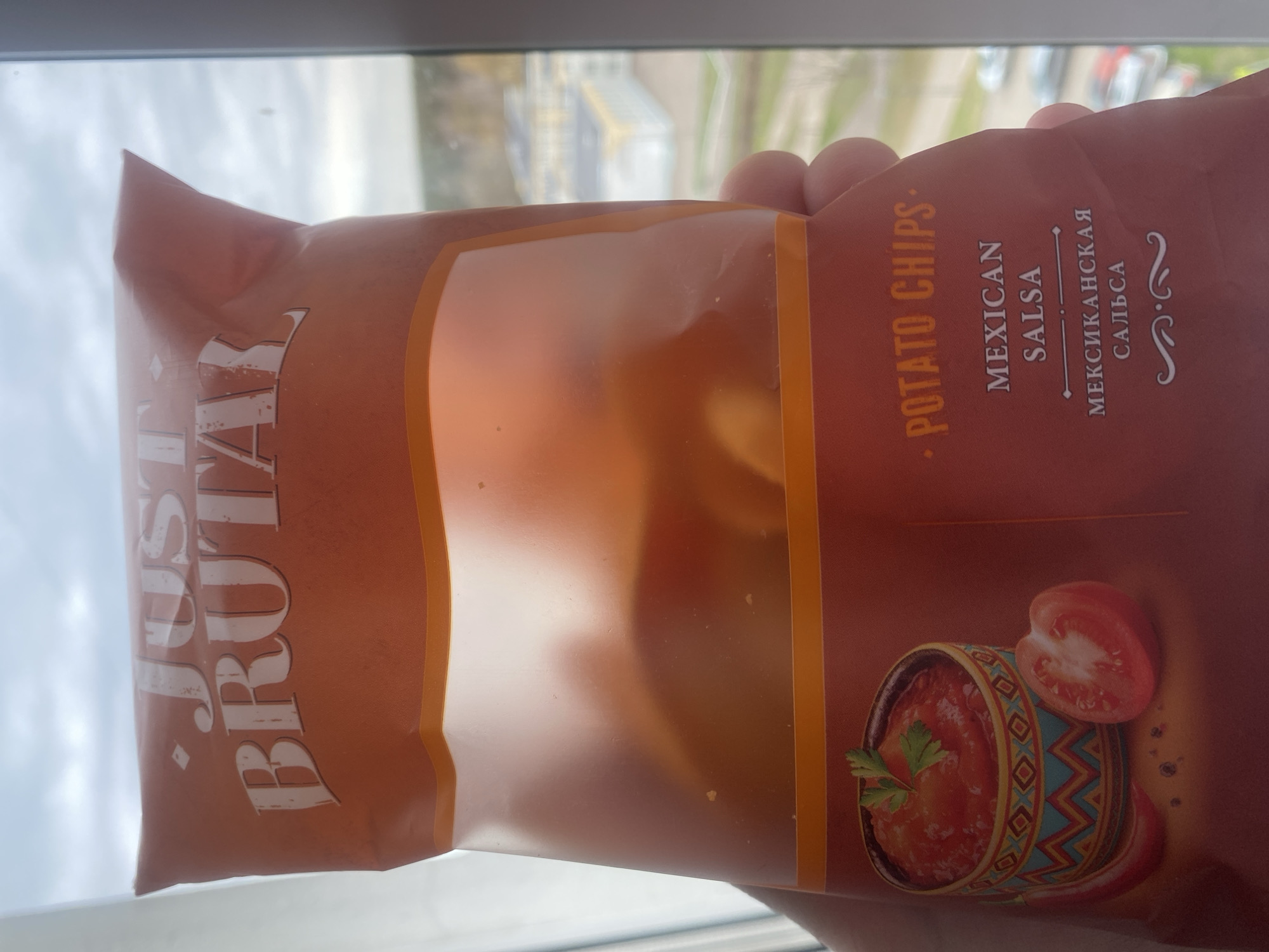 The image shows a bag of 'Just Brutal' potato chips, Mexican Salsa flavor. The bag is mostly brown and features a clear window showing the chips inside. The product name, flavor, and a picture of salsa with a tomato are visible on the bag. The bag is being held indoors, near a window that looks out onto a city, possibly in Unknown, Unknown.