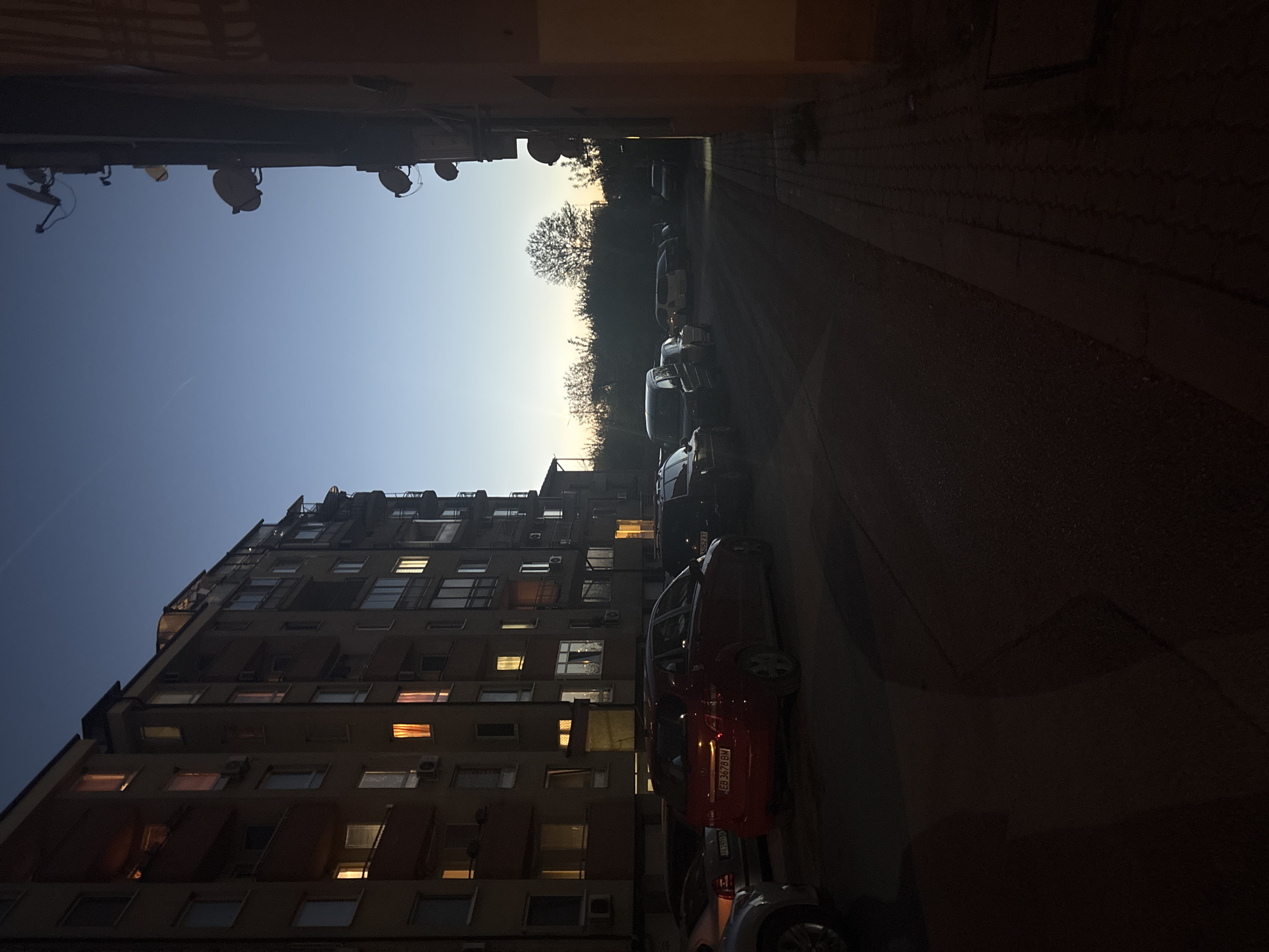 The image shows a street in Sofia, Bulgaria, lined with parked cars and a tall apartment building. The building appears to be residential with multiple floors and numerous windows, some of which are illuminated. The sky is visible between the buildings and at the end of the street, suggesting twilight or dawn. Satellite dishes are on the roof of the left building. The street is dimly lit, casting shadows and giving the scene a subdued atmosphere. There are no visible people.