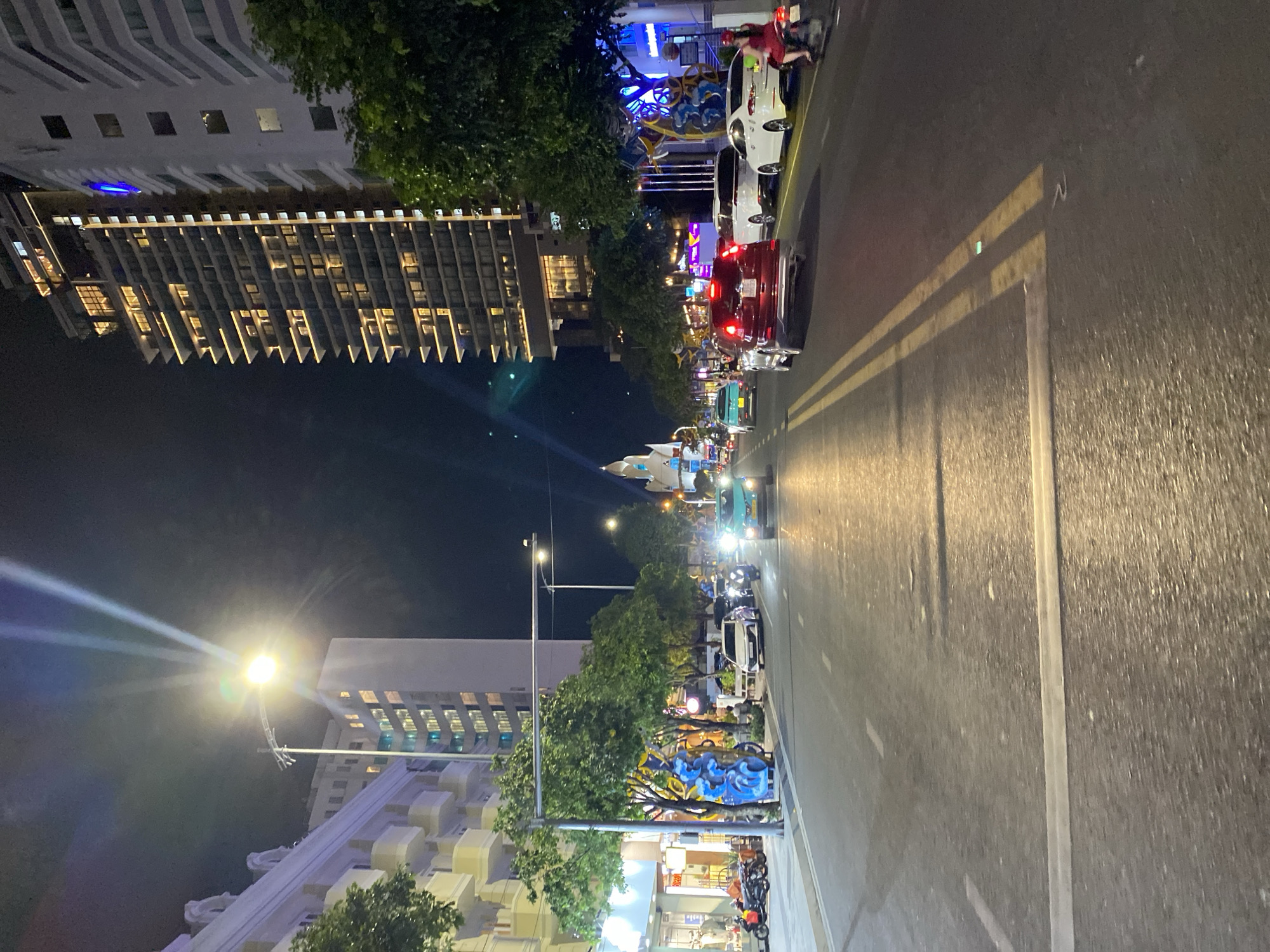The image depicts a street scene at night in Nha Trang Ward, Vietnam. The perspective is from a moving vehicle, looking down a wide, asphalt road illuminated by streetlights and the lights from buildings. On the left, a white building with balconies and a tree are visible. Across the street, a tall, modern building with many illuminated windows dominates the right side of the frame. The road has yellow double lines marking the lanes. Several vehicles, including cars and motorcycles, are present on the road, some with their headlights on, creating reflections on the wet pavement. Pedestrians are not clearly visible, but a person on a motorcycle in a red dress is seen on the right side of the road. The overall atmosphere is one of a bustling urban environment at night, with traffic and illuminated structures.