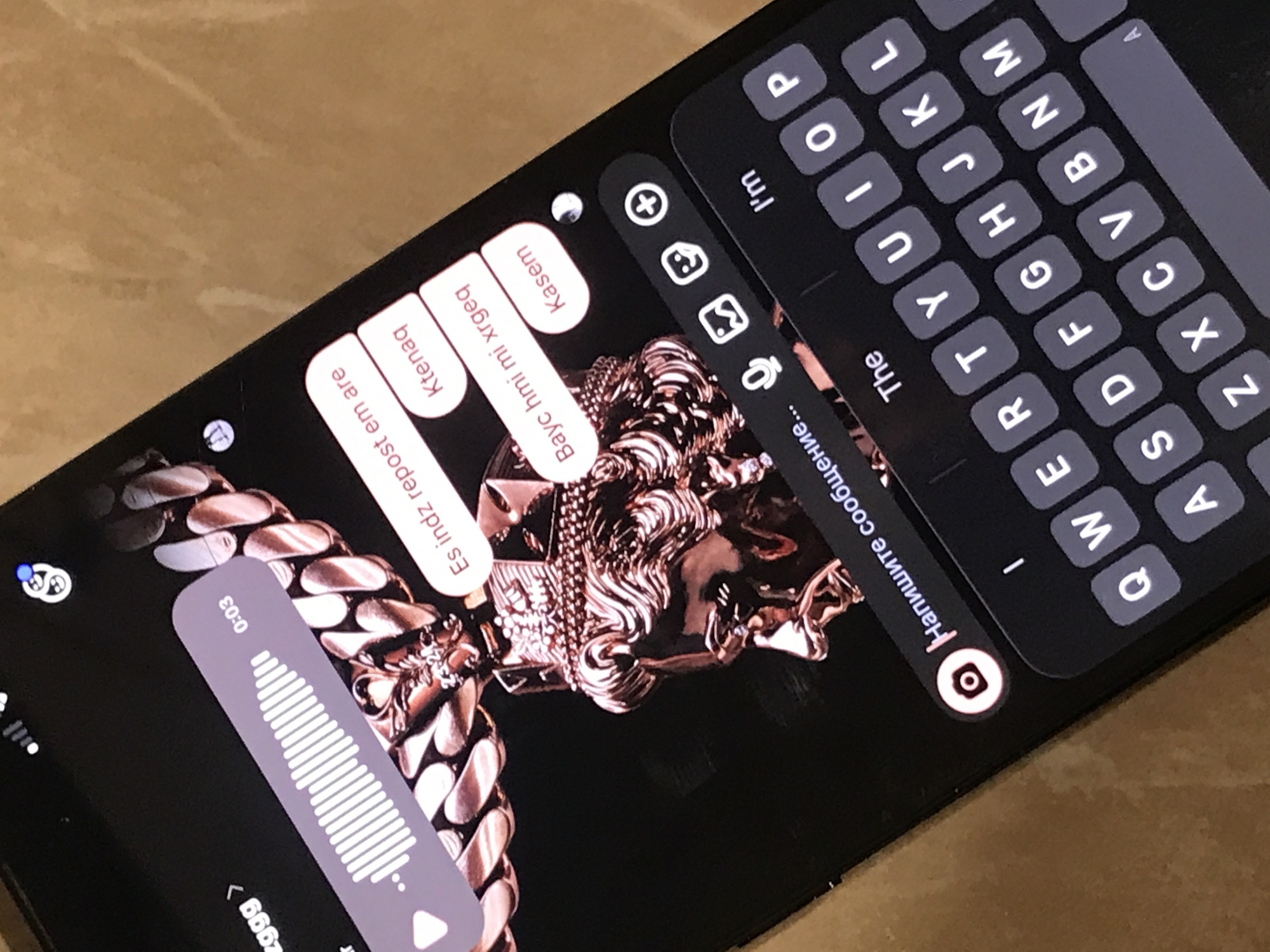 The image shows a smartphone displaying a messaging application. The screen displays suggested replies in Armenian along with the English word "Kasem" which is used in Armenia too. The keyboard is visible, indicating an active text input field in Russian. A music player interface is present in the bottom left corner. The background wallpaper is a close-up of a gold chain and pendant. The phone is laying on a table, which appears to be made of wood. There is no indication that this is in Yerevan, Armenia.