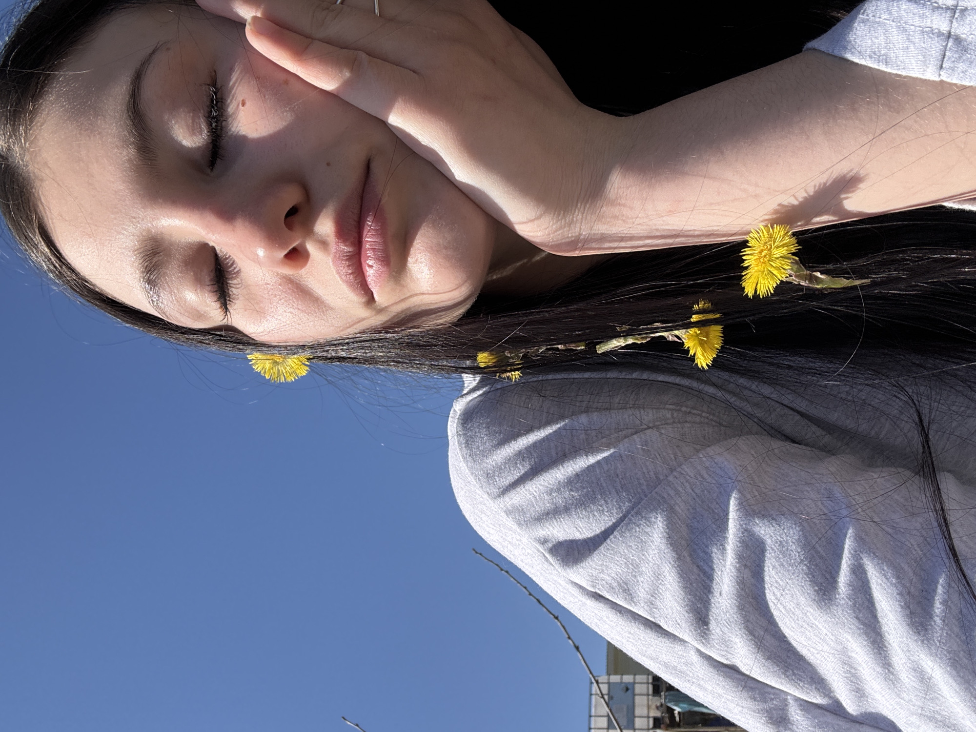 A woman with long, dark hair is angled against a clear, blue sky. She is wearing a light gray long-sleeved shirt. Her eyes are closed, and she is touching her face with her hand. There are several small yellow flowers interspersed in her hair. The background includes a hint of a building exterior with tiled walls. The location is unknown and cannot be confirmed as the city of Фоки, Russia, based on the given image.