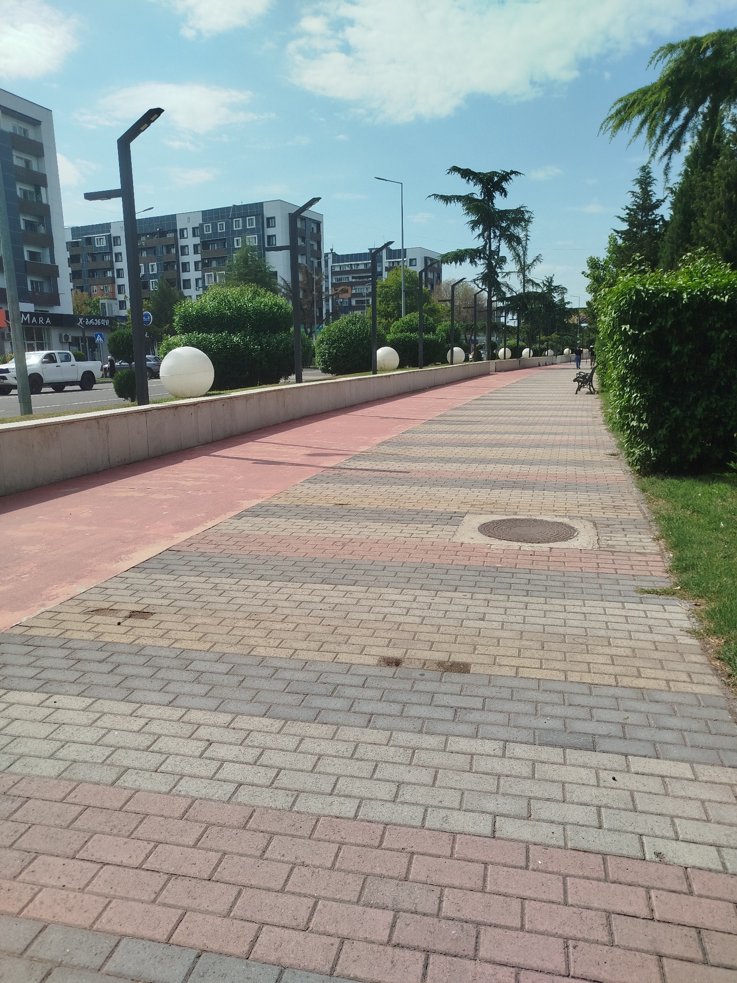 The image depicts a paved pedestrian walkway with a red-colored bicycle lane adjacent to it. The walkway is made of interlocking pavers in a repeating pattern of pink, grey, and beige. Several spherical white decorative spheres are positioned along a low concrete wall that separates the walkway from the road and buildings. Beyond the wall, modern residential buildings with multiple stories and balconies are visible, along with manicured green bushes and trees. Streetlights with modern designs are also present. On the right side of the walkway, a grassy area and a dense hedge are visible. A dark-colored bench is situated near the hedge, and a single person is walking away from the camera on the walkway. A circular metal manhole cover is embedded in the pavement in the foreground. The scene appears to be in an urban setting on a sunny day with a partly cloudy sky. This scene is located on Rustaveli Avenue in Rustavi, Georgia.